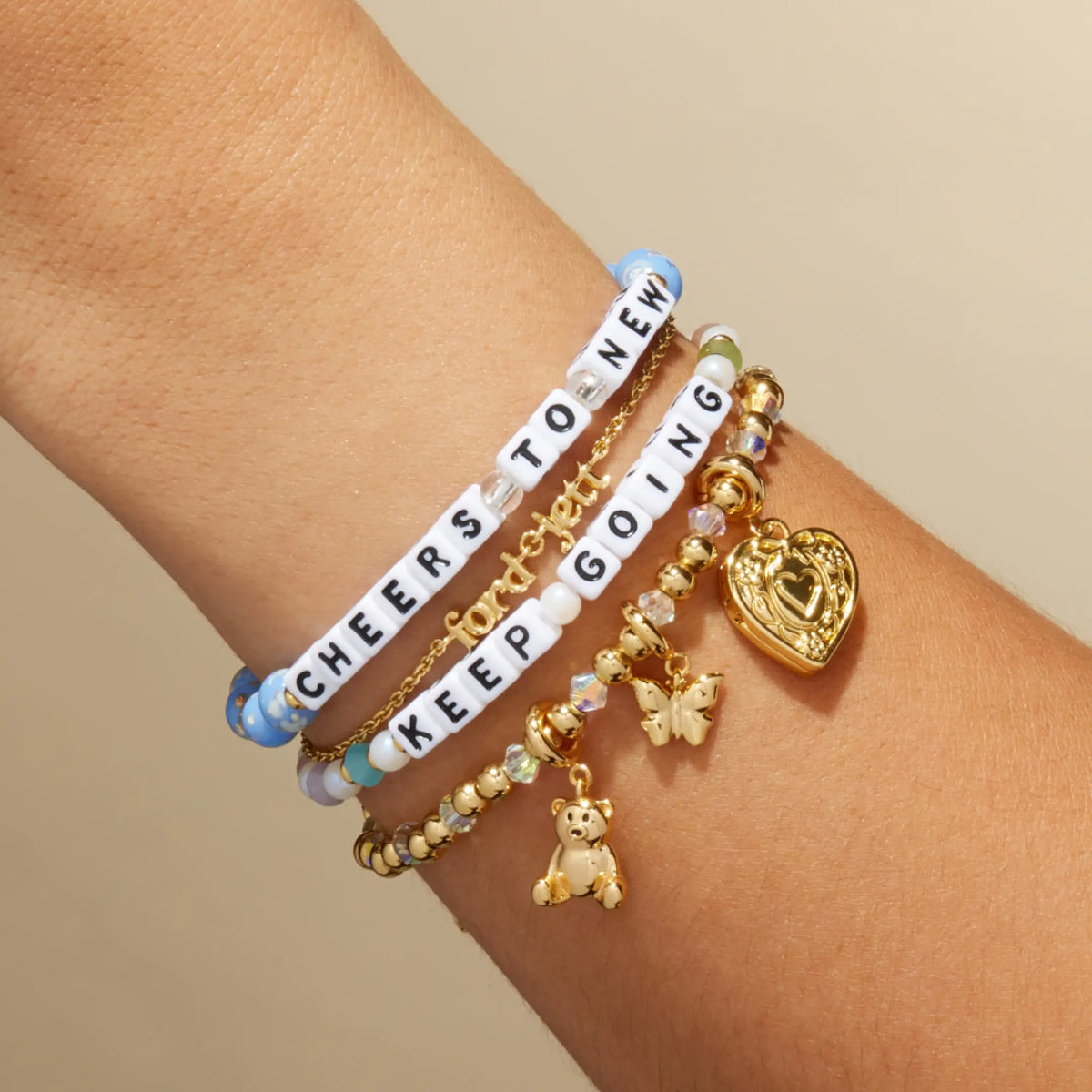 Custom Glacier Glow bracelet with white letter beads reading Cheers To New stacked with a Keep Going bracelet and gold charm bracelets, on-model on wrist against a tan background