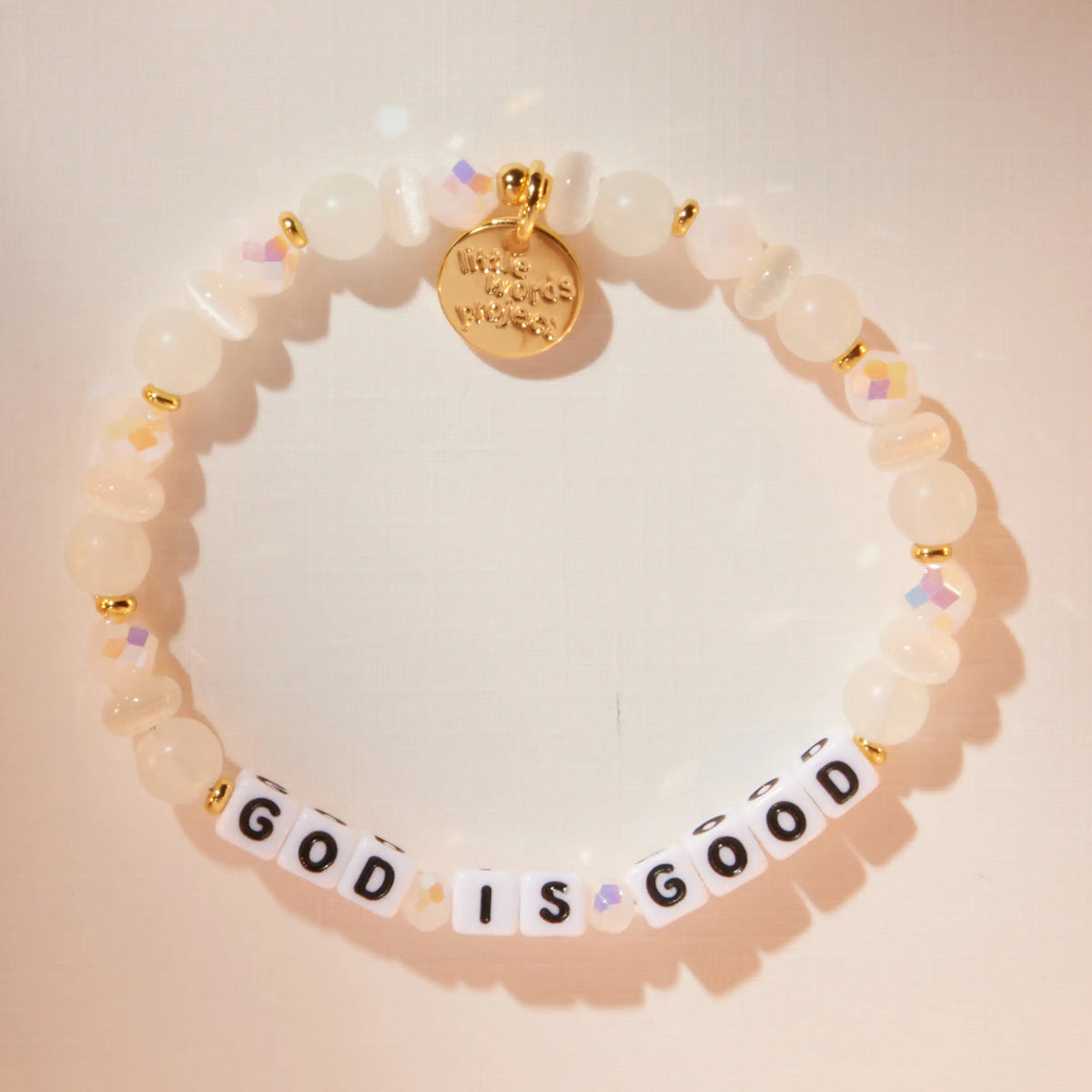 God is Good bracelet in the Luxe Knit bead pattern with white letter beads, flat-lay on warm neutral background with soft light