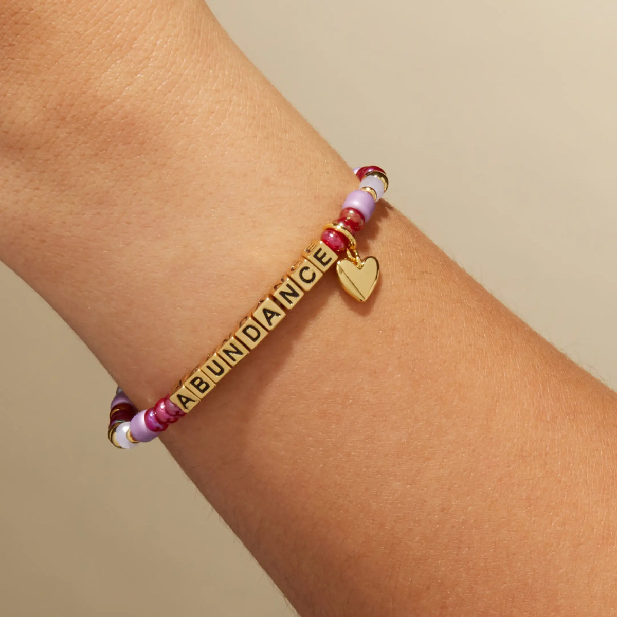 Custom Golden Cherry bracelet with gold letter beads reading Abundance, cranberry red and mauve beads with a gold heart charm, on-model on wrist against a tan background