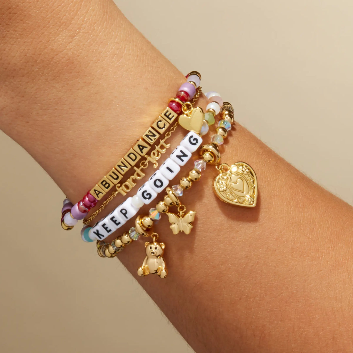 Custom Golden Cherry bracelet with gold letter beads reading Abundance stacked with a Keep Going bracelet and gold charm bracelets, on-model on wrist against a tan background