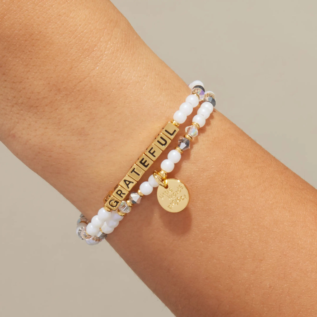Gold Grateful bracelet in the Shiny Things bead pattern with gold letter beads, stacked with a coordinating beaded bracelet, on-model on wrist against cream background