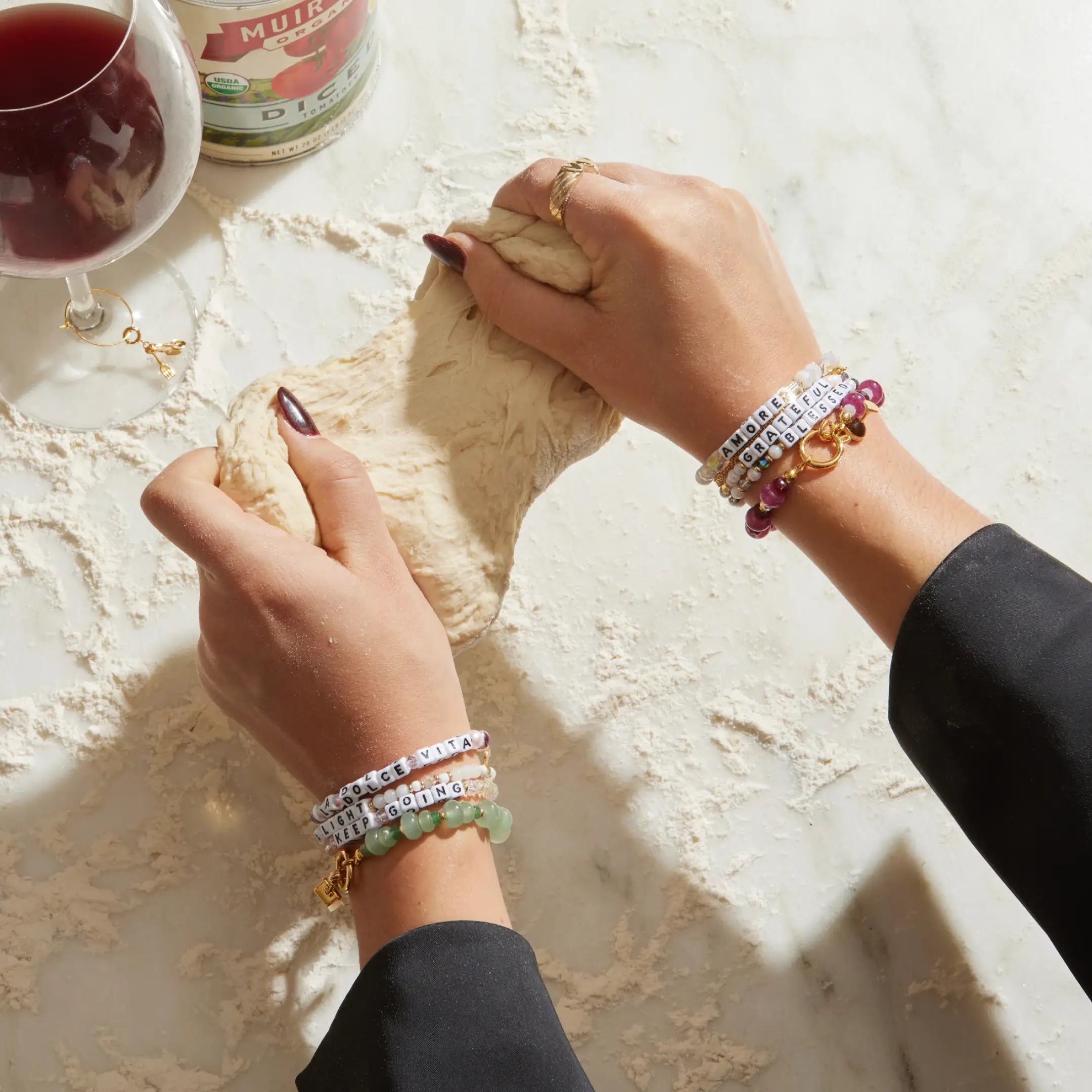 Person rolling dough with bracelets on a light surface, next to a glass of red wine and a can.