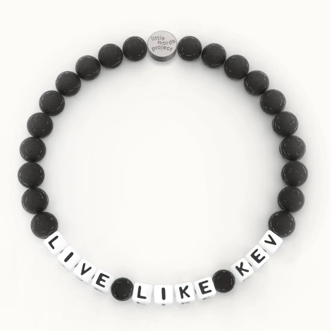 Live Like Kev giveback bracelet in the Lava Stone bead pattern, white letter beads with black lava stone beads and a silver logo tag, flat-lay on cream background