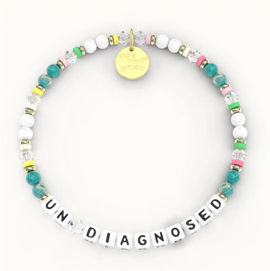 Un Diagnosed giveback bracelet in the Joyful bead pattern, white letter beads with teal stone, white howlite and clear crystal beads and pink, green and yellow spacers with gold accents, flat-lay on cream background