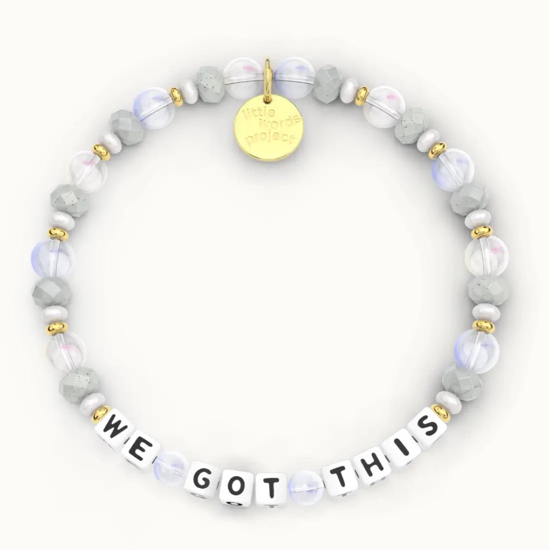 We Got This giveback bracelet in the Calm Waters bead pattern, white letter beads with gray stone, frosted clear and iridescent glass beads with gold accents, flat-lay on cream background
