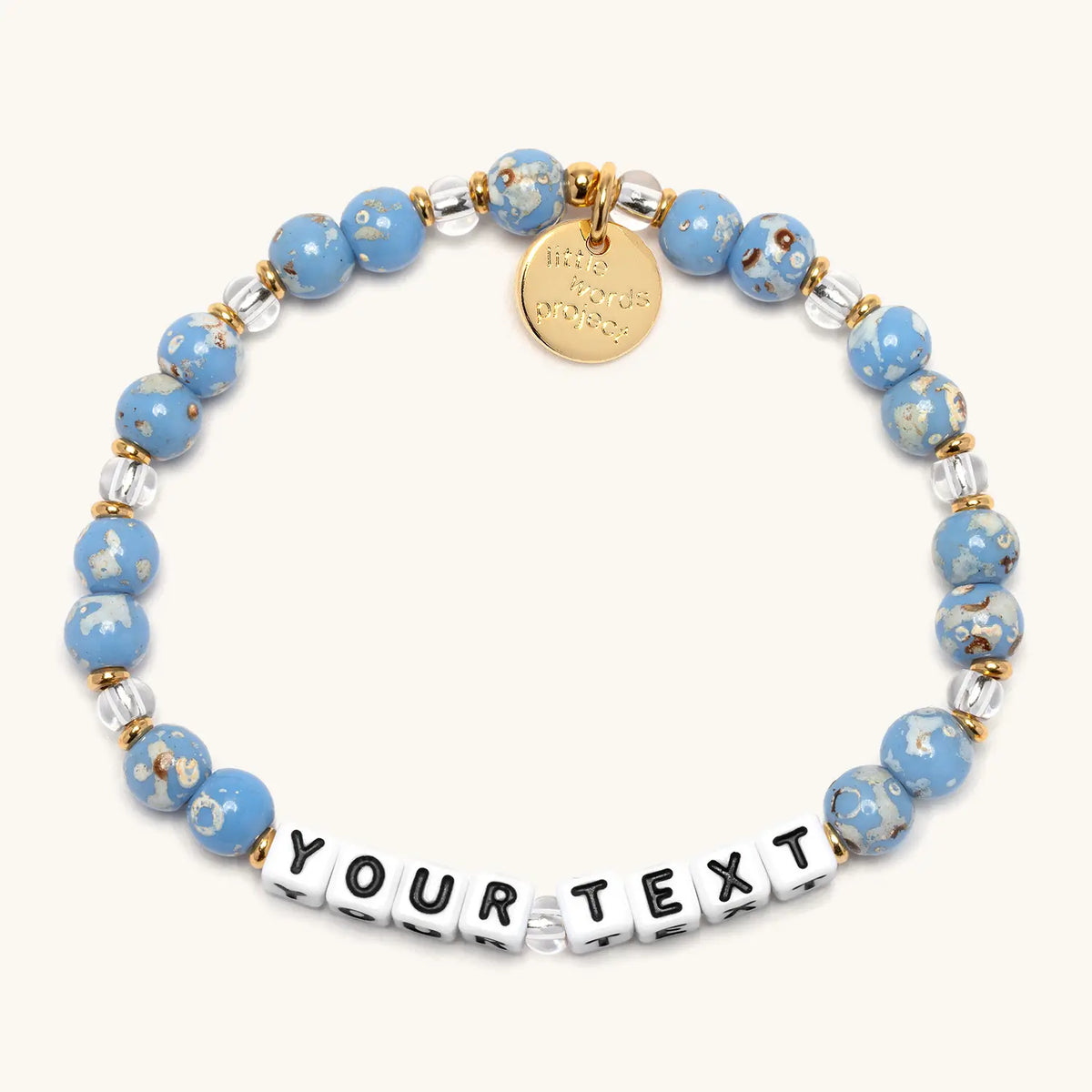 Custom Glacier Glow bracelet in the Glacier Glow bead pattern, white letter beads reading Your Text, light blue speckled beads and clear crystal beads with gold accents, flat-lay on cream background