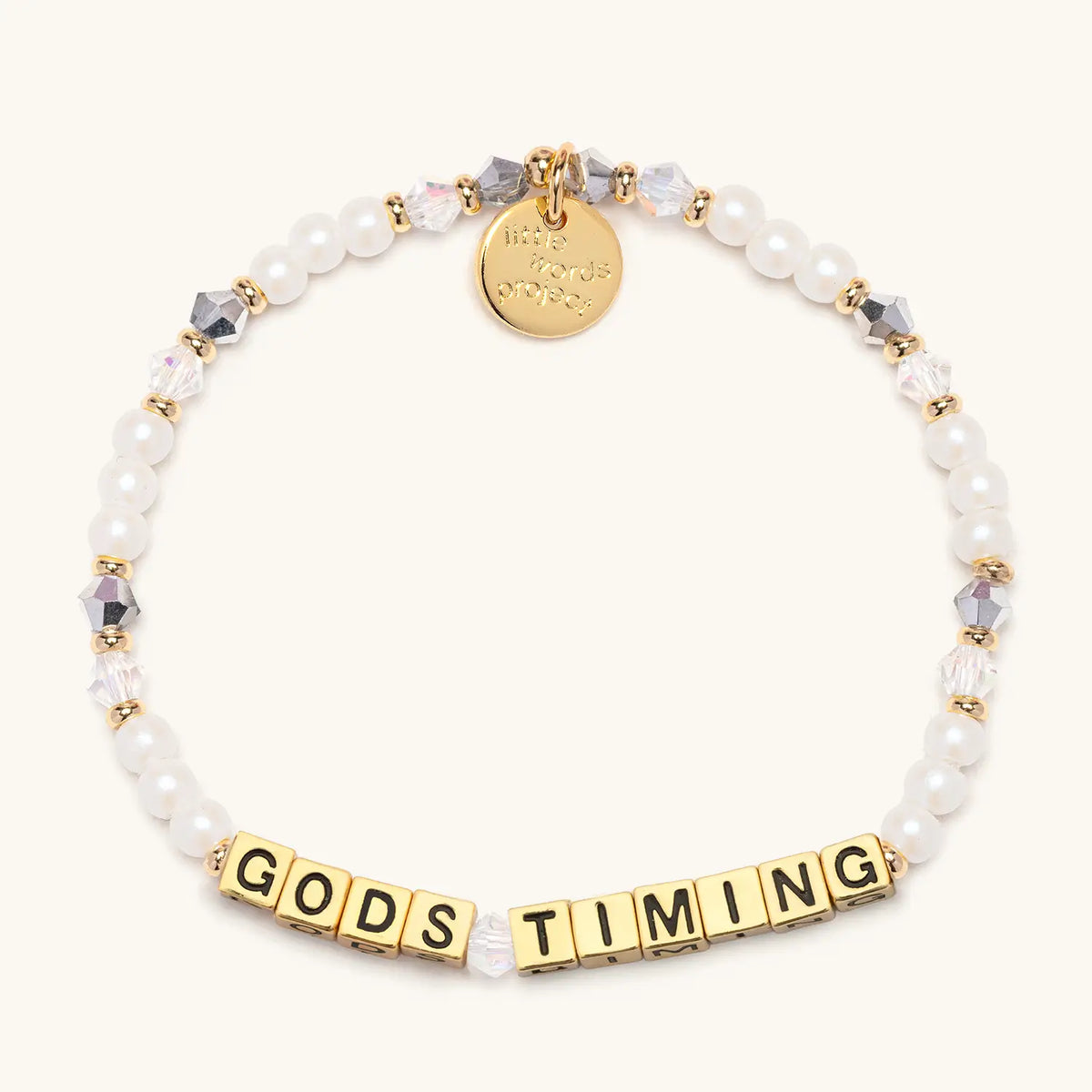 God's Timing bracelet with gold letter beads, white pearls, clear and silver crystal beads, gold spacers and a gold Little Words Project charm, flat-lay on a cream background