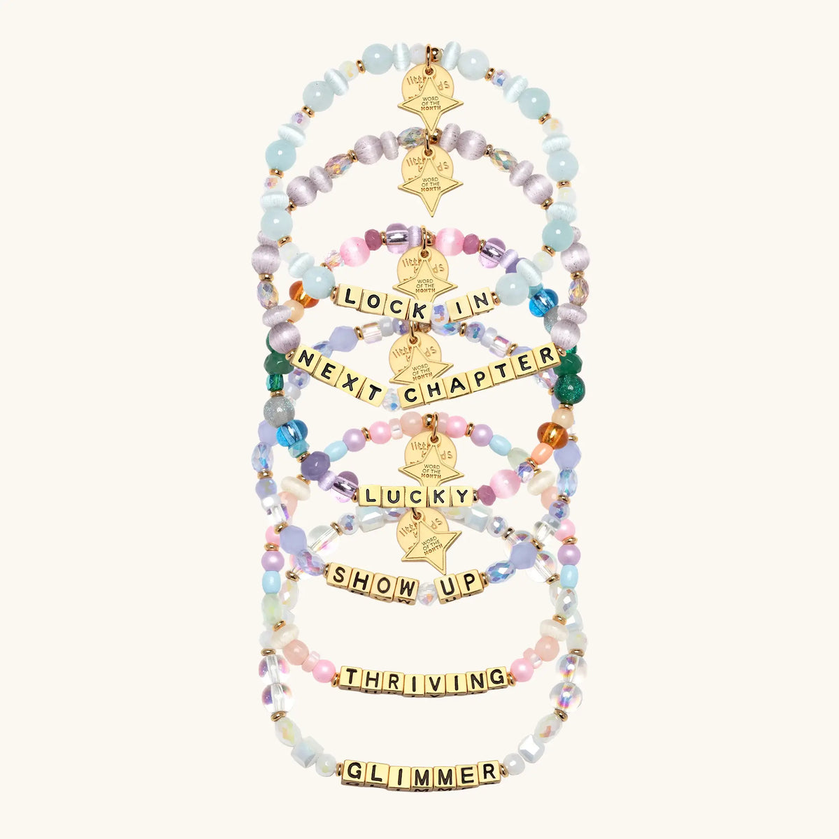 Six beaded stretch bracelets arranged vertically on a light cream background. Each bracelet features pastel-colored beads, gold-toned letter blocks spelling motivational phrases like 'LOCK IN', 'NEXT CHAPTER', 'LUCKIY', 'SHOW UP', 'THRIVING', and 'GLIMMER', and gold star tags.