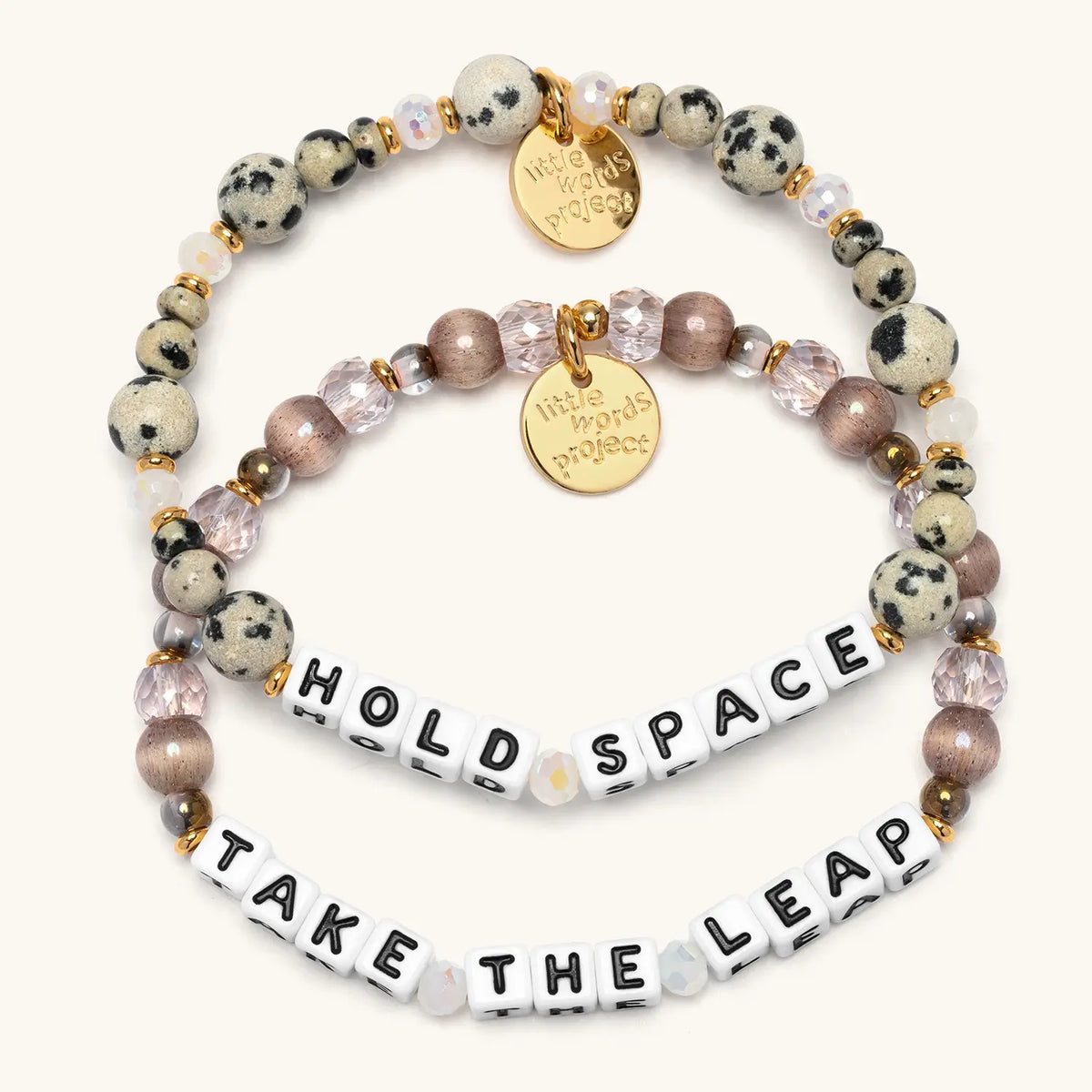 Take the Leap Bracelet Set flat-lay on cream background showing two stacked bracelets: the Hold Space bracelet in white letter beads with dalmatian jasper stone beads and iridescent white crystals, and the Take the Leap bracelet in white letter beads with smoky mauve cat eye beads, blush pink faceted crystals, and metallic bronze beads, both with gold accents