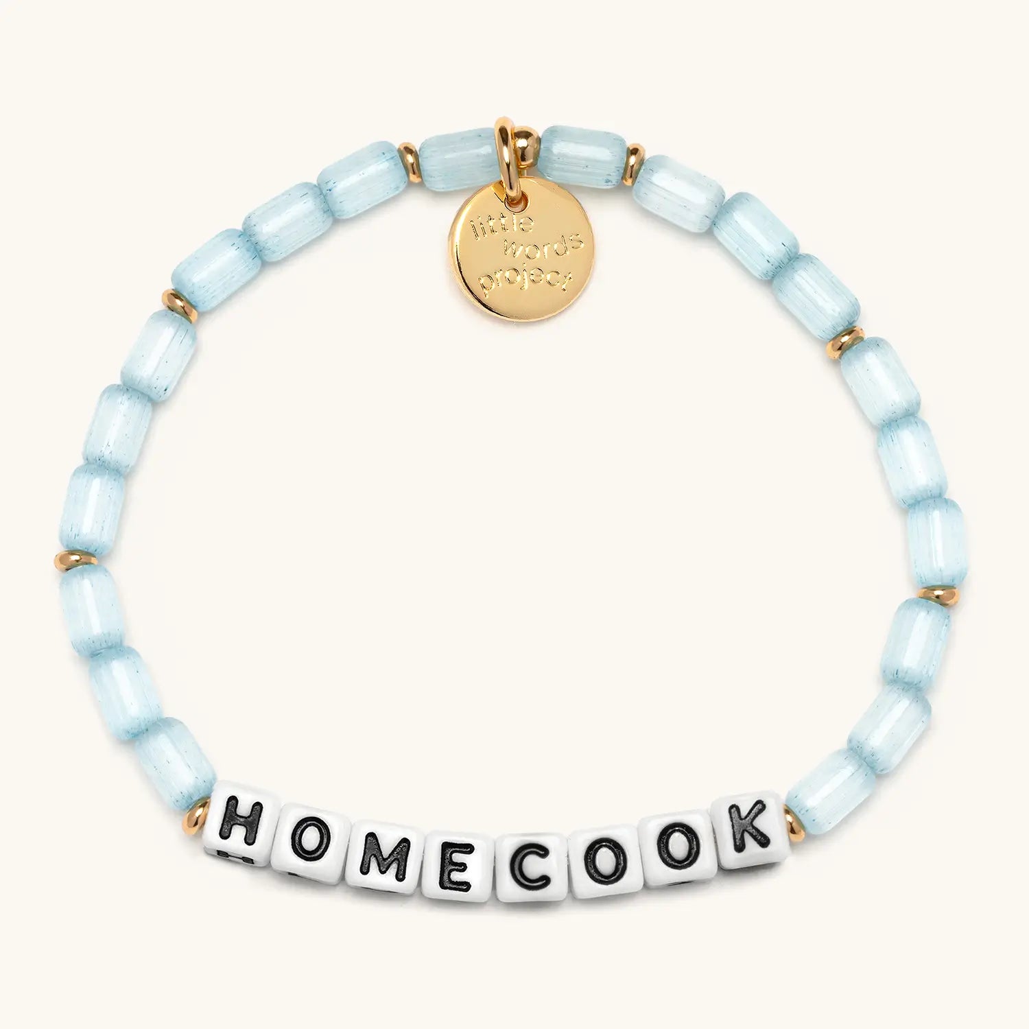 Homecook bracelet in Sunday Blues from the Little Words Project x Alex Snodgrass collaboration, with pale blue glass beads, gold spacers, and white letter beads spelling HOMECOOK, finished with a gold Little Words Project charm tag.
