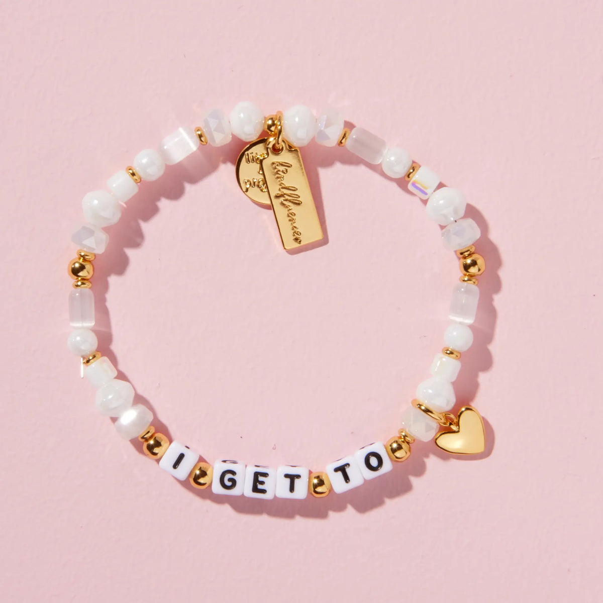 I Get To Charm Bracelet in the Ivory Sparkle bead pattern, white letter beads with white faux pearls, frosted glass, and iridescent crystal beads with gold accents, a gold heart charm and gold Kindfluencer tag, flat-lay on pink background