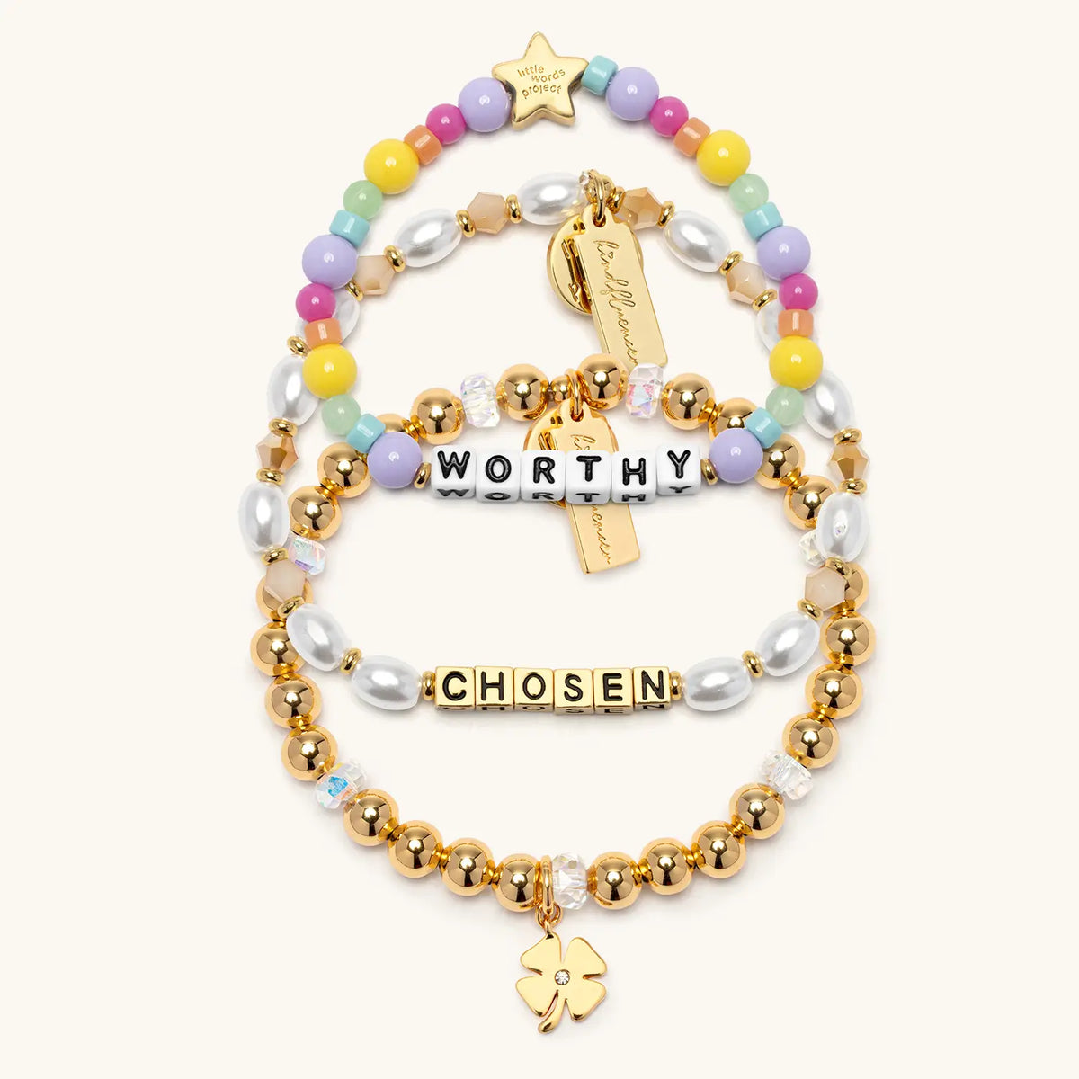 Balance Bracelet Set by Kirstin Czernek flat-lay on cream background showing three stacked bracelets: the Chosen bracelet with white faux pearls, the gold beaded Four Leaf Clover Charm Bracelet, and the Worthy kids bracelet with pastel rainbow beads and a gold star