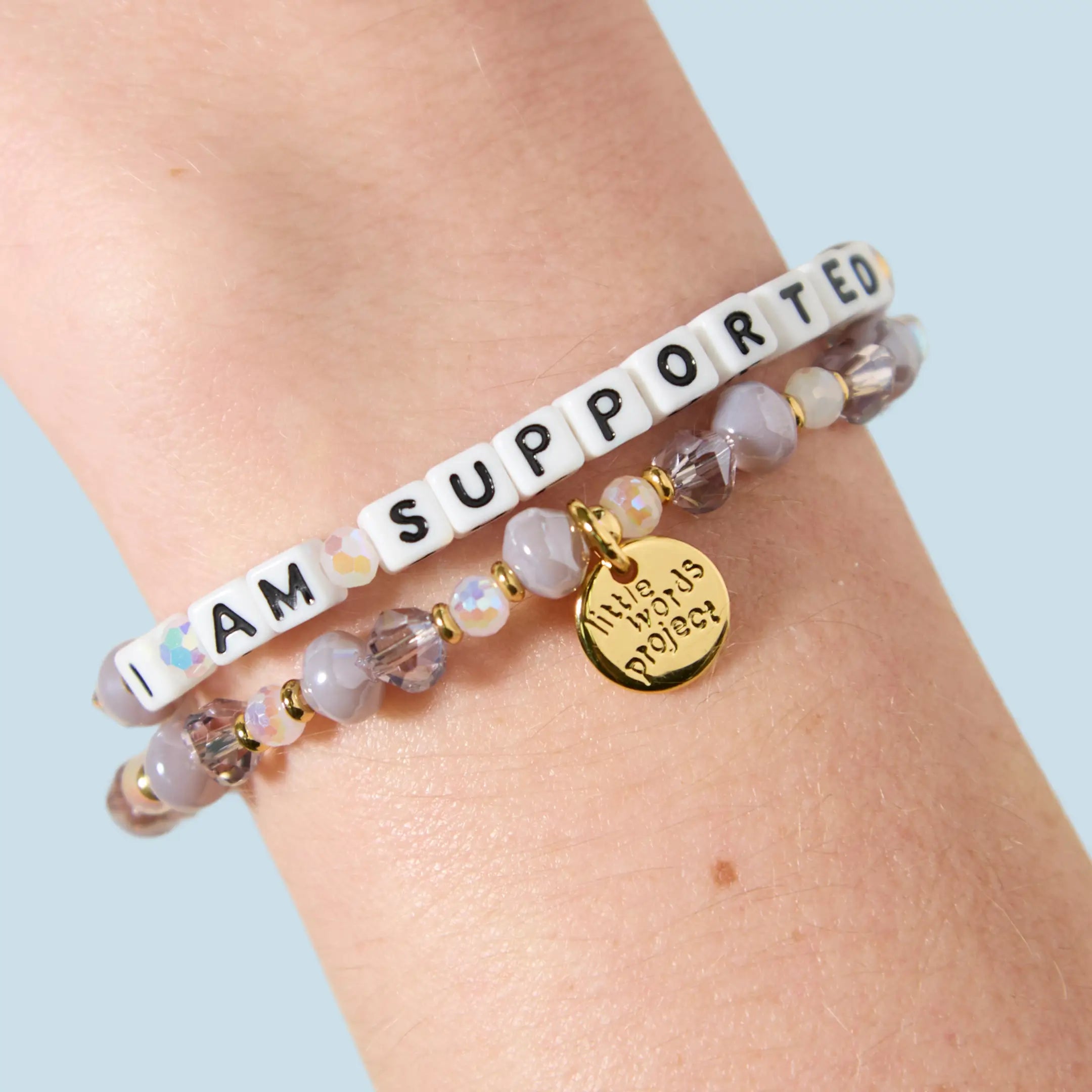 Two bracelets stacked on a wrist — one with white letter beads reading "I Am Supported" and one with grey, mauve, and iridescent crystal beads featuring a gold Little Words Project charm pendant.