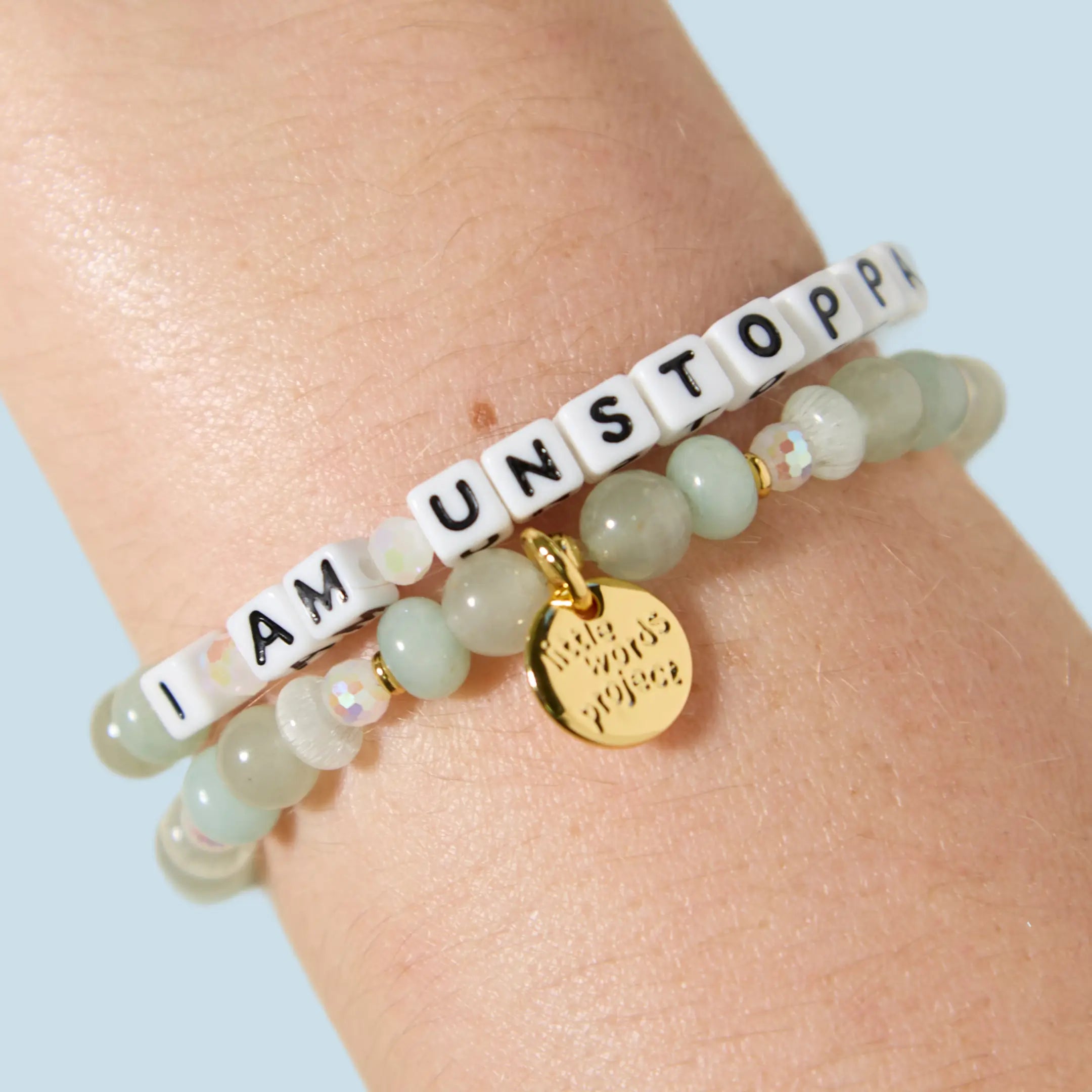 Two bracelets stacked on a wrist — one with white letter beads reading "I Am Unstoppa[ble]" and one with mint green and iridescent beads featuring a gold Little Words Project charm pendant.