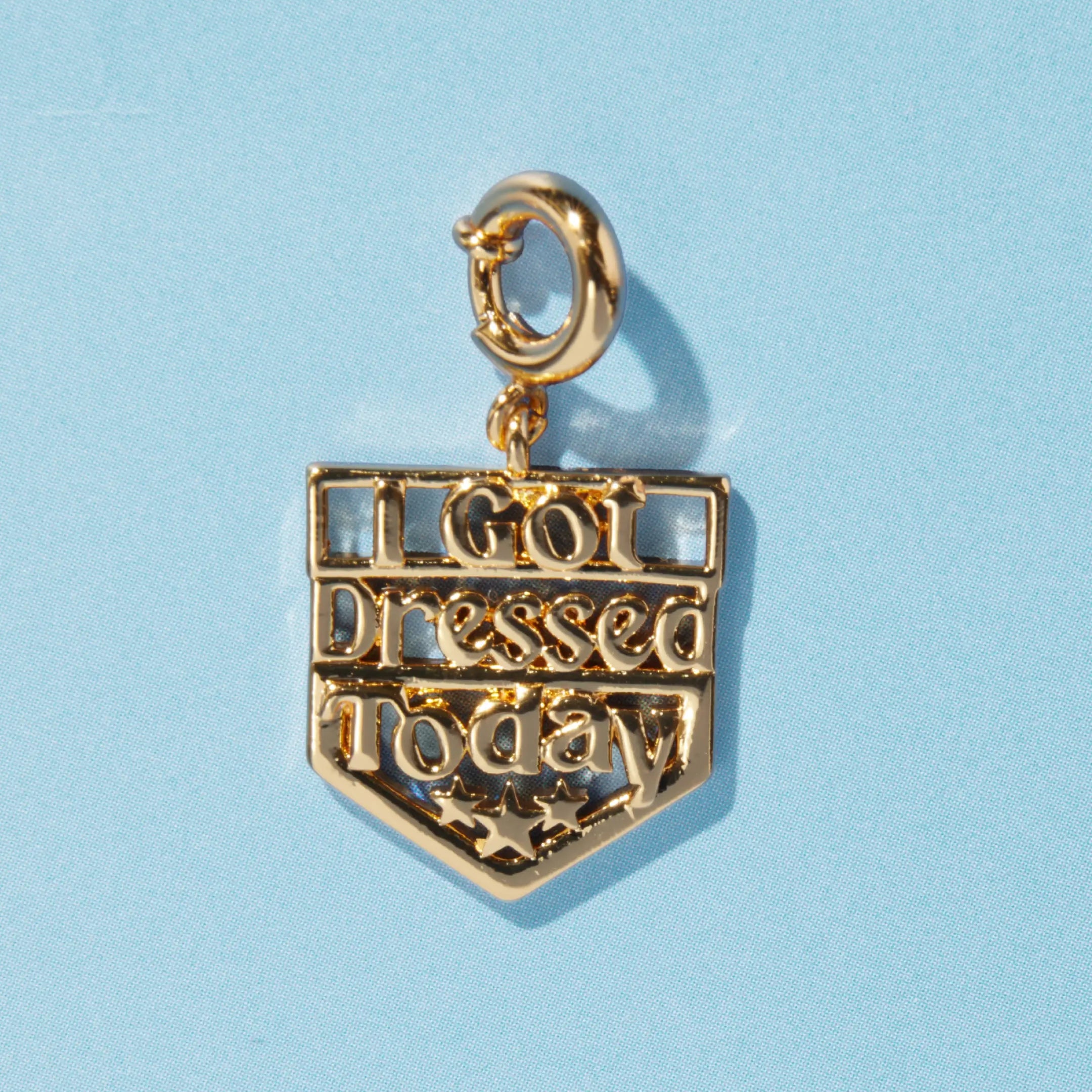 A gold shield-shaped charm reading "I Got Dressed Today" with decorative stars at the bottom, attached to a spring ring clasp, on a blue background.