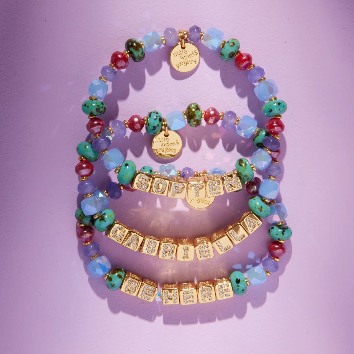Three BOLDER Custom Jewel Tones bracelets with gold CZ letter blocks, teal speckled, plum, burgundy, and periwinkle faceted beads with gold LWP charms, flat-lay on lavender background