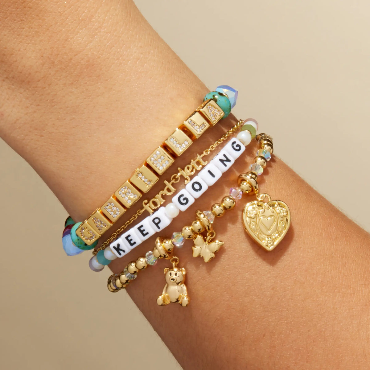 BOLDER Custom Jewel Tones bracelet with gold CZ letter blocks stacked with a Keep Going bracelet and gold charm bracelets, on-model on wrist against a tan background