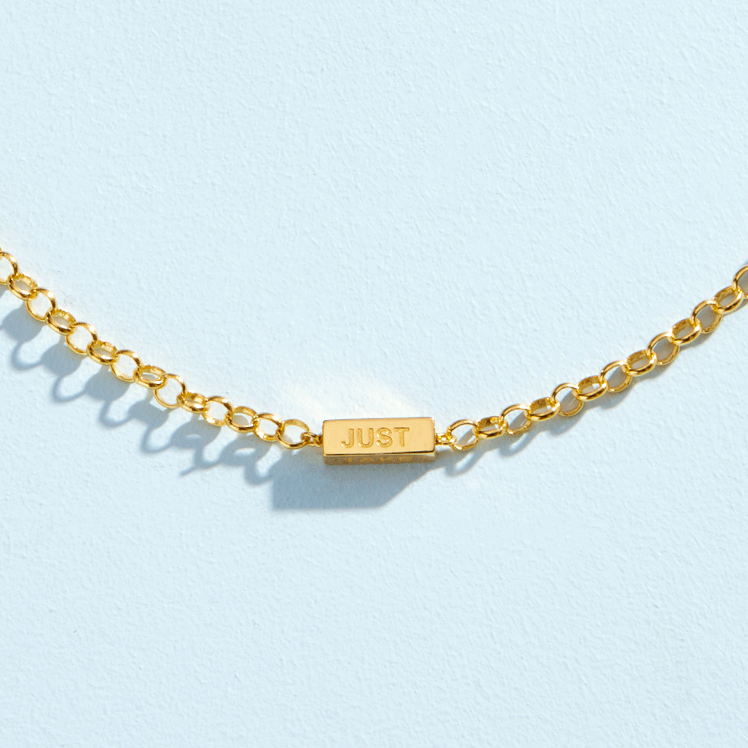 Close-up of a gold chain bracelet with an engraved "JUST" bar charm on a light blue background.