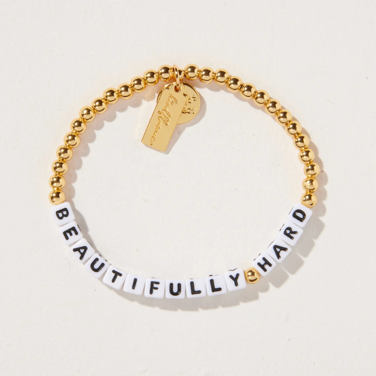Beautifully Hard bracelet by Kirstin Czernek, white letter beads with gold plated beads and gold accents, detail flat-lay on light background