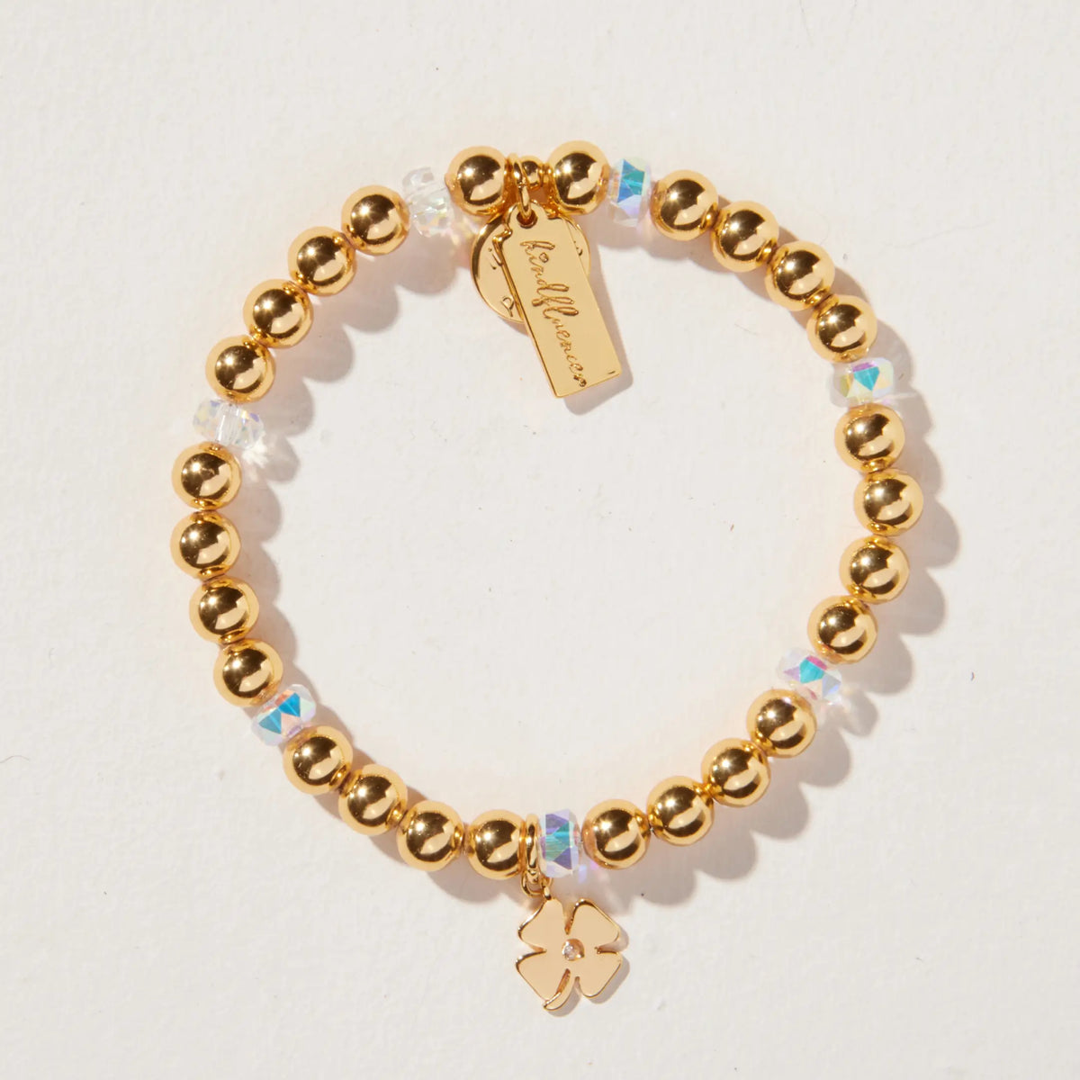 Four Leaf Clover Charm Bracelet by Kirstin Czernek, gold plated beads with clear iridescent crystals and a gold four leaf clover charm, detail flat-lay on light background