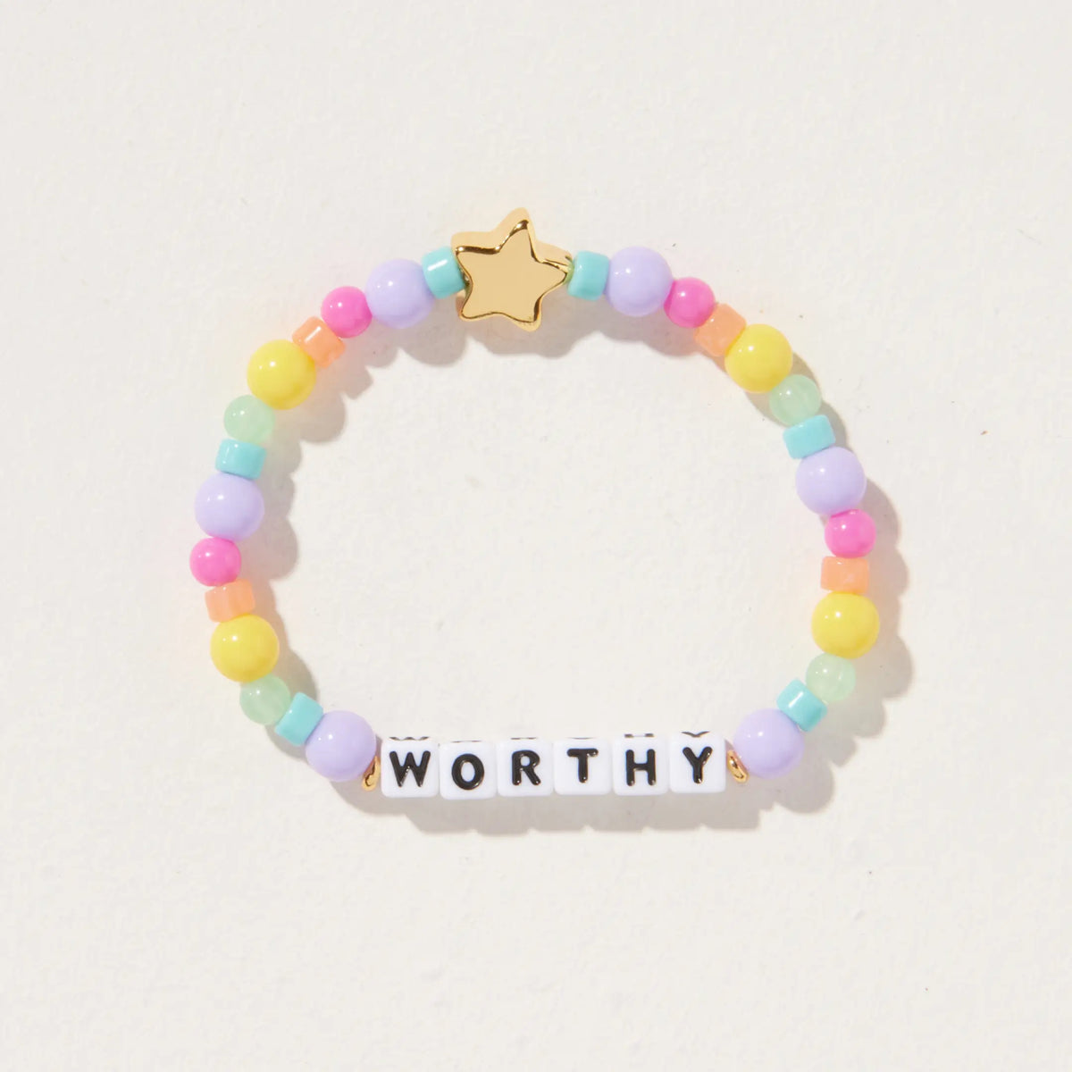 Worthy kids bracelet by Kirstin Czernek, white letter beads with pastel rainbow beads and a gold star bead, detail flat-lay on light background