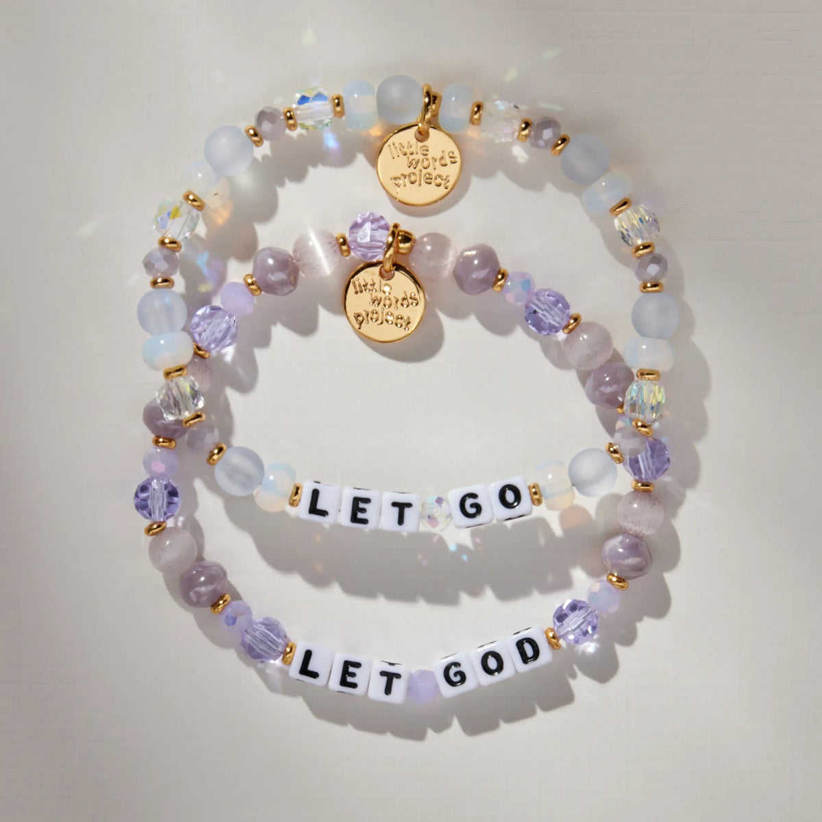 Let Go Let God Bracelet Set flat-lay showing the Let Go bracelet in frosted light blue and opal beads and the Let God bracelet in lavender crystals and mauve cat eye beads, both with white letter beads