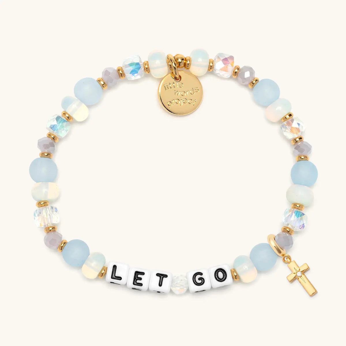 Let Go bracelet in the Marshmellow bead pattern with white letter beads, gold puffy cross charm with a clear CZ stone, flat-lay on cream background