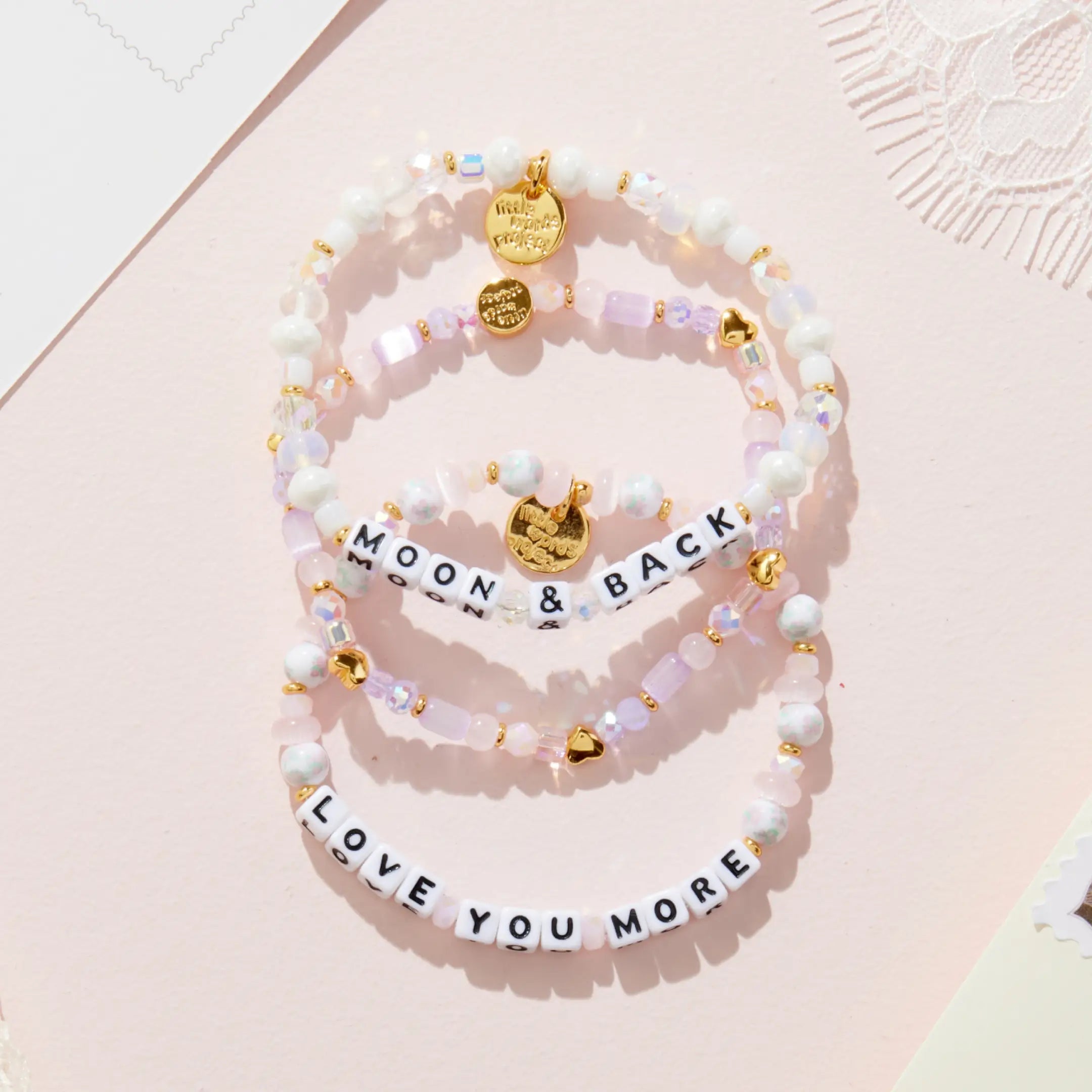 Set of beaded bracelets with 'Moon & Back' and 'Love You More' on a light pink background