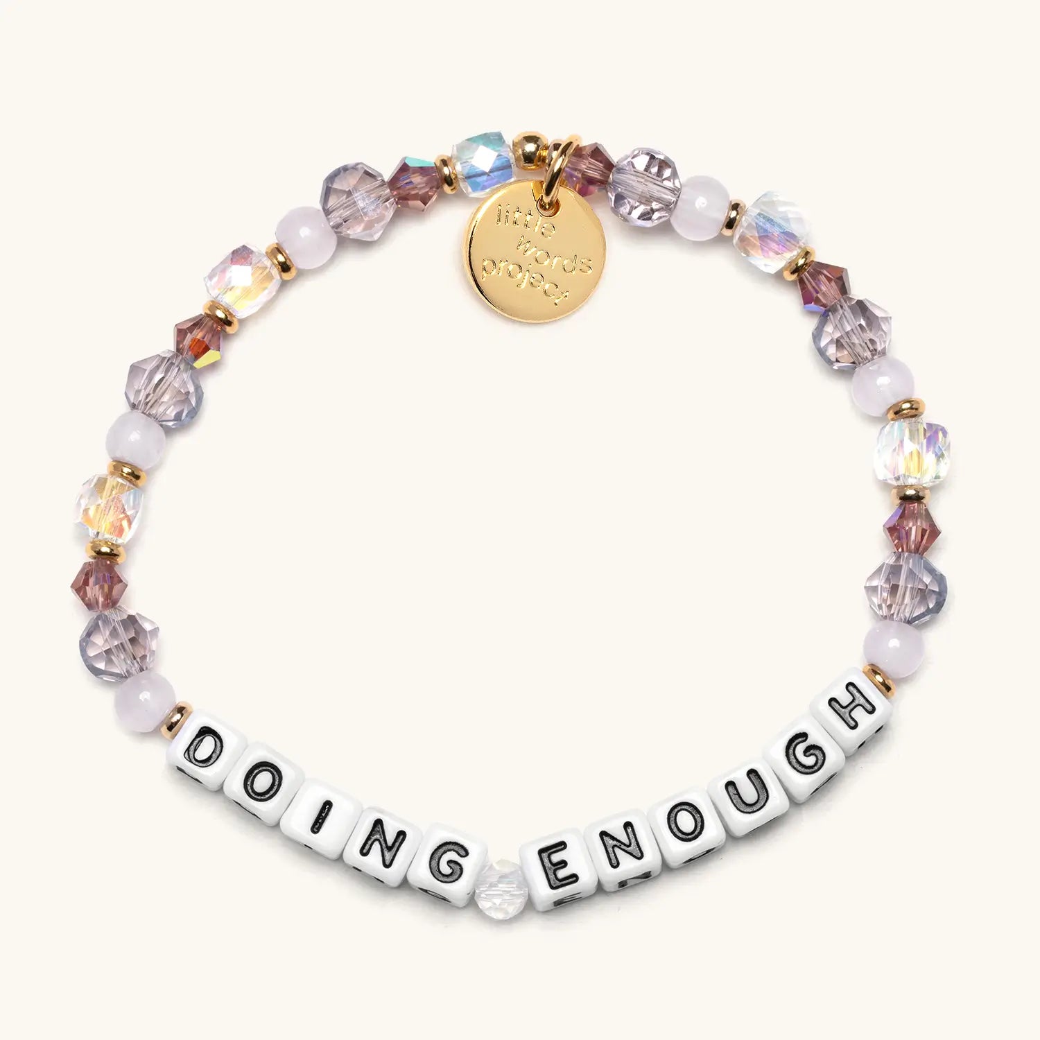 A beaded stretch bracelet featuring white square letter beads spelling "DOING ENOUGH," interspersed with iridescent crystal and pink/purple faceted beads and small gold accent spacers. A round gold charm engraved with a logo hangs from the bracelet. The overall color palette is soft lavender, blush, and gold on a light neutral background.
