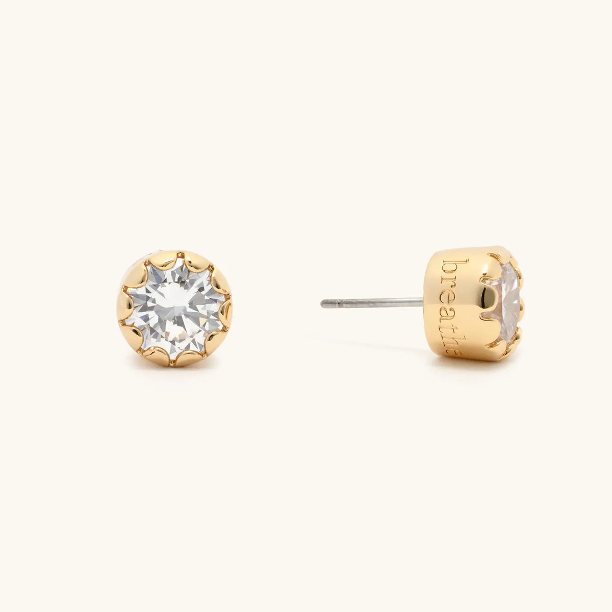 Breathe Scalloped Bezel Studs, 18k gold-plated studs with a clear CZ stone in a scalloped bezel and breathe engraved on the side, flat-lay on cream background