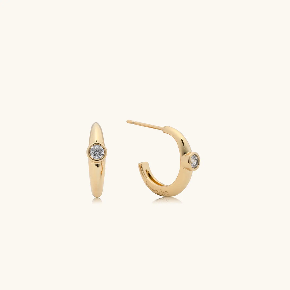 Breathe Puffy Huggies, 18k gold-plated puffy huggie earrings with a round clear CZ stone and breathe engraved inside the hoop, flat-lay on cream background