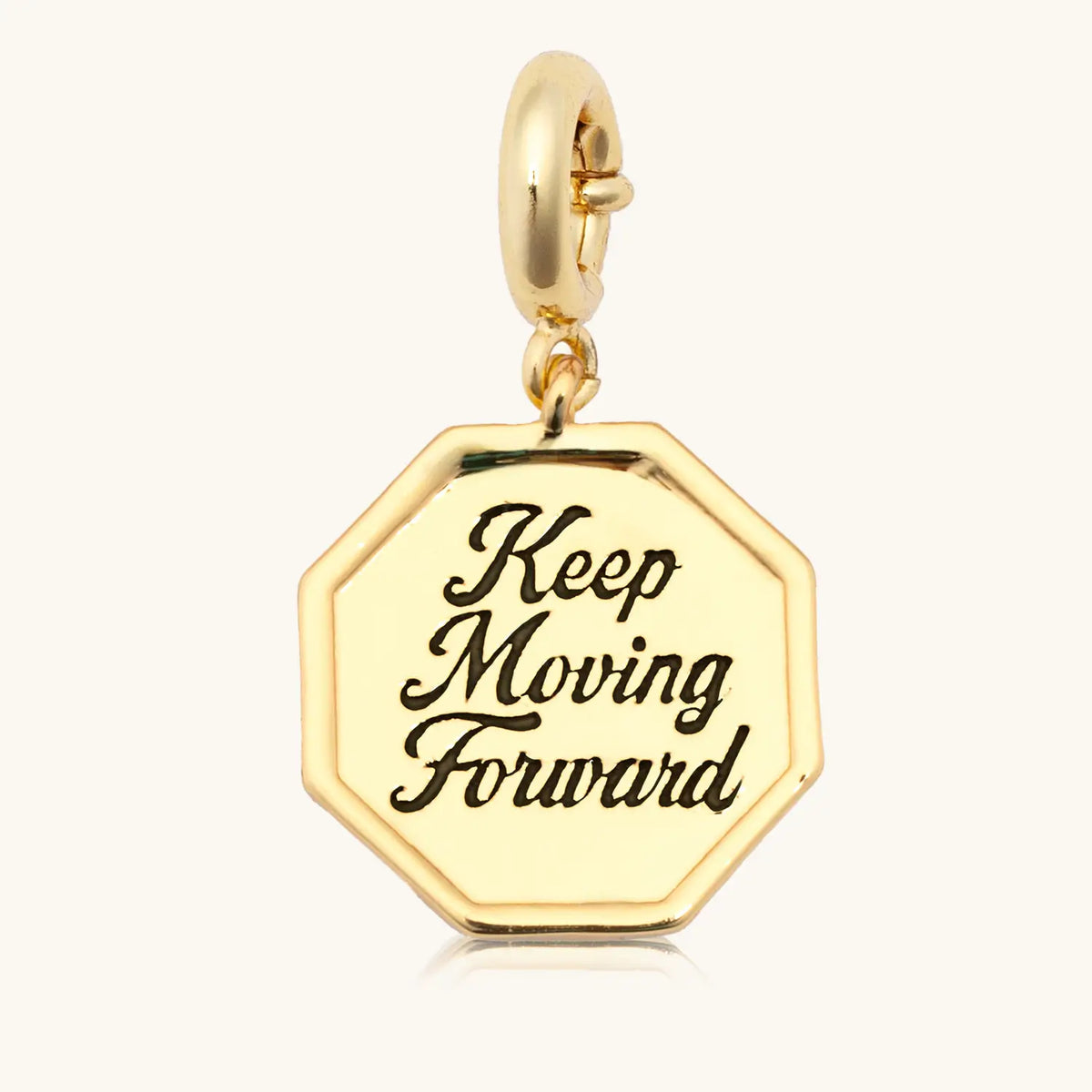 Keep Moving Forward Charm front, 18k gold-plated octagon medallion engraved Keep Moving Forward in script on a gold spring ring clasp, detail on cream background