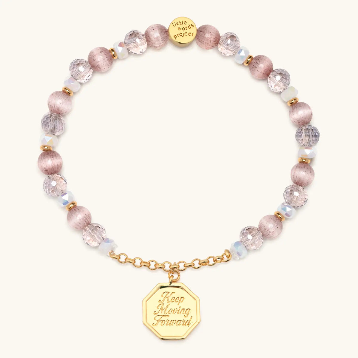 Keep Moving Forward Charm Bracelet, pink cat eye, mauve crystal, and clear glass beads with gold chain and an octagon medallion engraved Keep Moving Forward, flat-lay on cream background