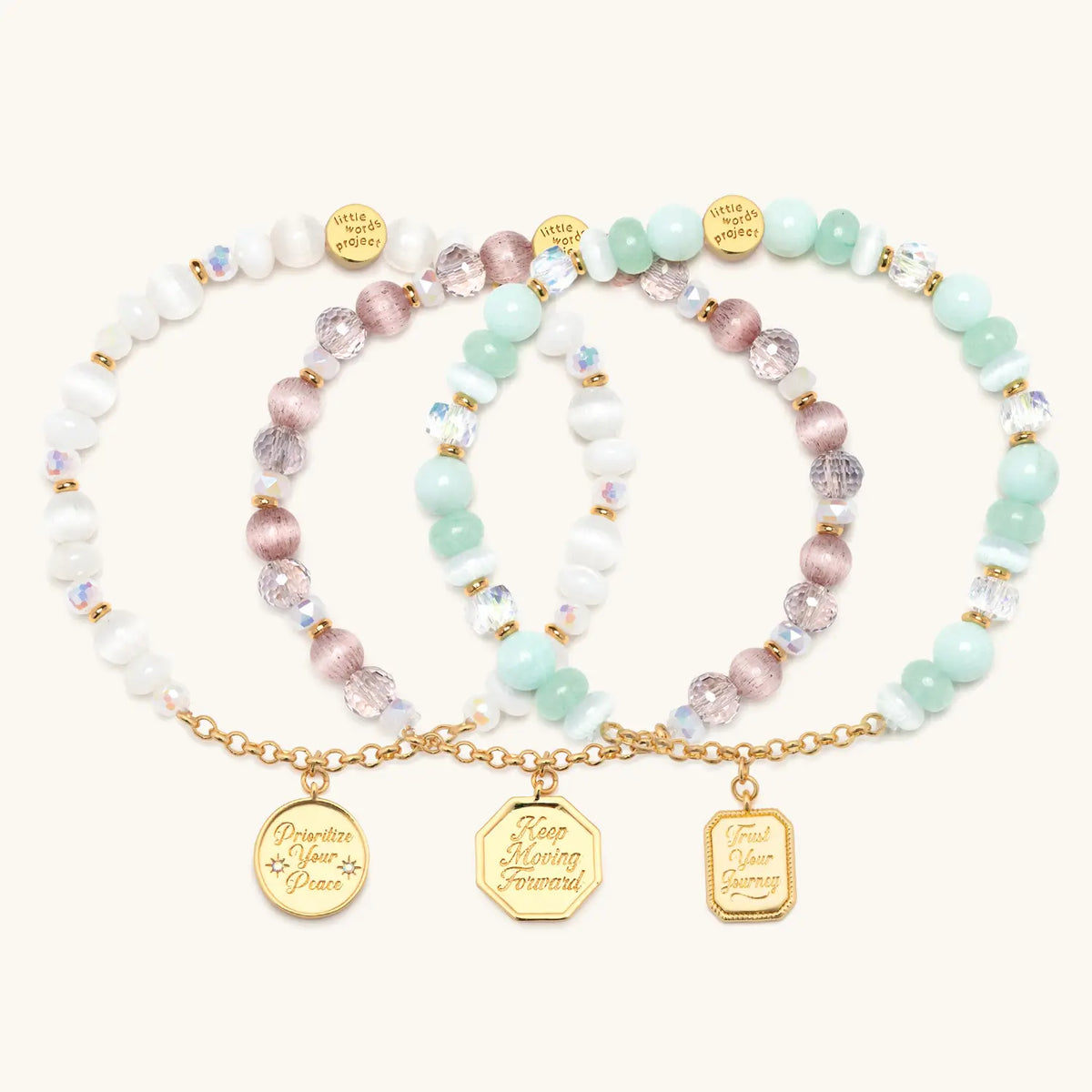 Prioritizing Me Bracelet Set flat-lay on cream background showing three stacked charm bracelets: Prioritize Your Peace in white cat eye beads, Keep Moving Forward in pink cat eye beads, and Trust Your Journey in seafoam beads, each with a gold medallion