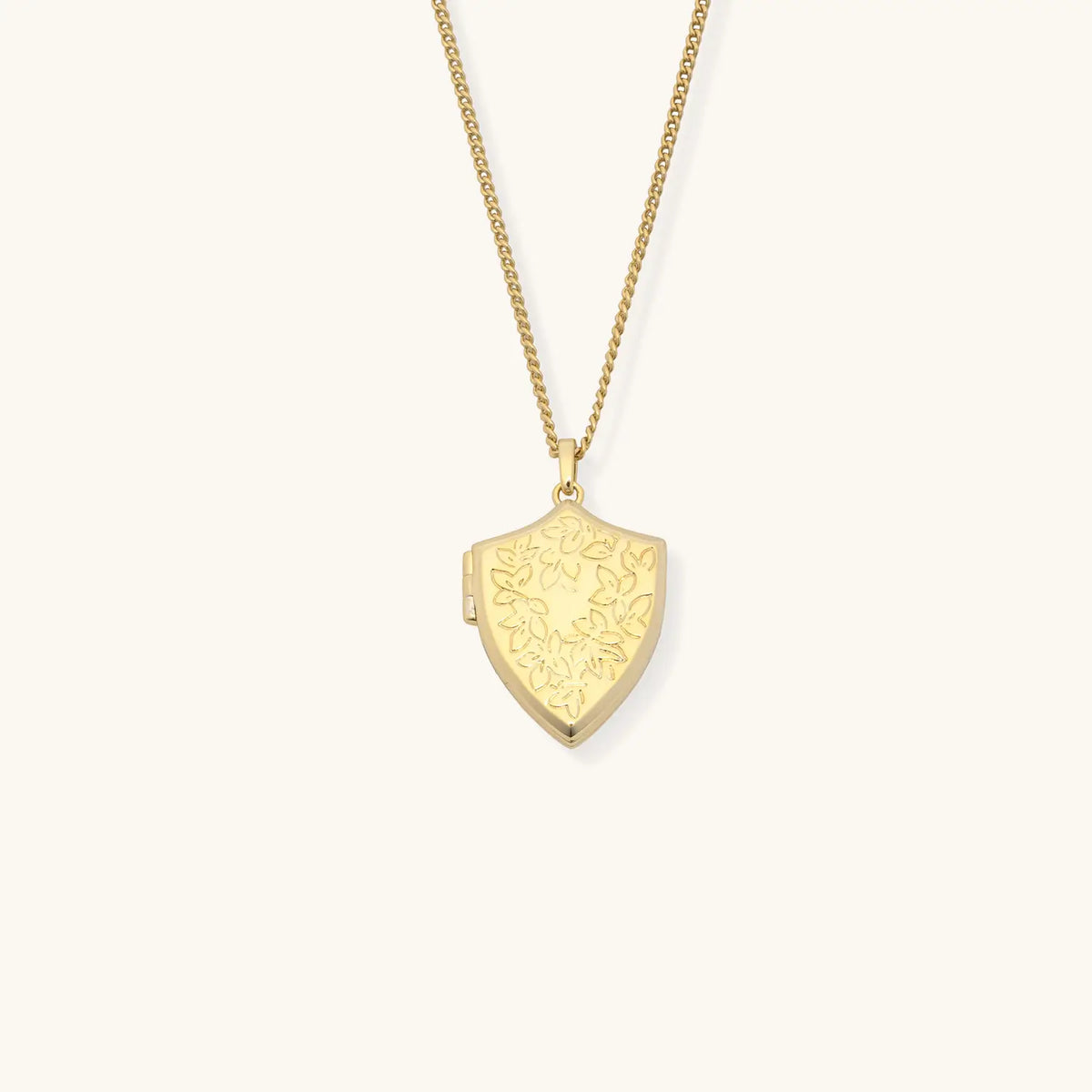 Shield Locket Necklace, 18k gold-plated shield locket with floral engraving on a gold curb chain, detail on cream background