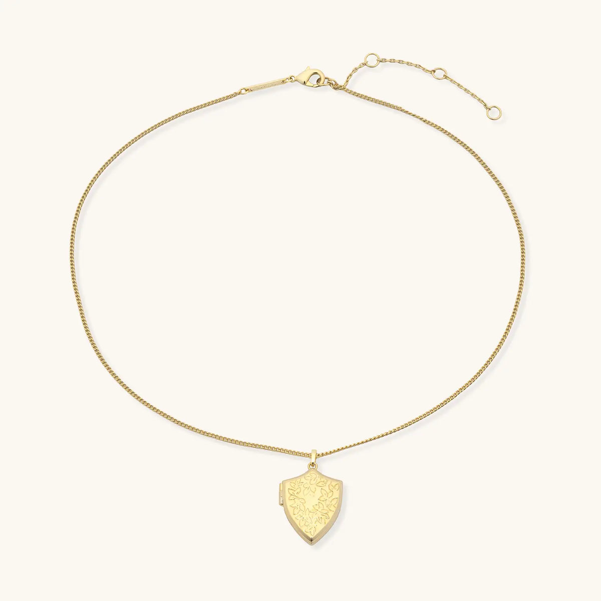 Shield Locket Necklace, full view of the 18k gold-plated shield locket on a gold curb chain with extender, flat-lay on cream background