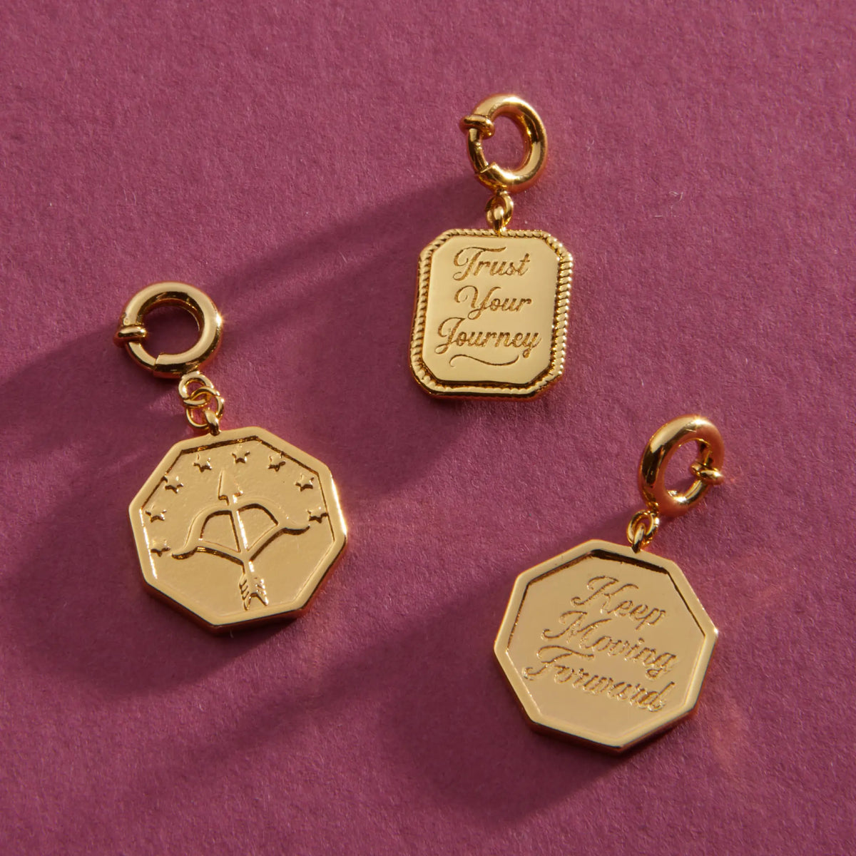 Keep Moving Forward and Trust Your Journey Charms, 18k gold-plated octagon and rectangular medallions with spring ring clasps, flat-lay on a berry background