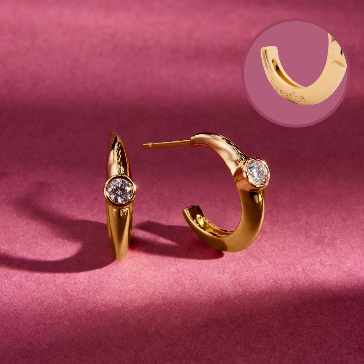 Breathe Puffy Huggies, 18k gold-plated huggie earrings with a round clear CZ stone, with an inset showing breathe engraved inside the hoop, detail on a berry background
