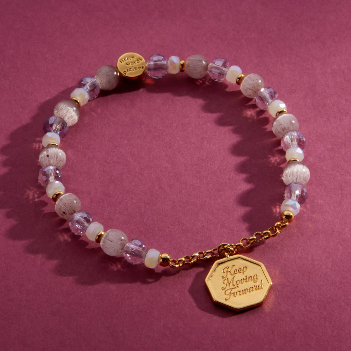 Keep Moving Forward Charm Bracelet, pink cat eye, lilac crystal, and clear glass beads with gold chain and an octagon medallion engraved Keep Moving Forward, flat-lay on a berry background