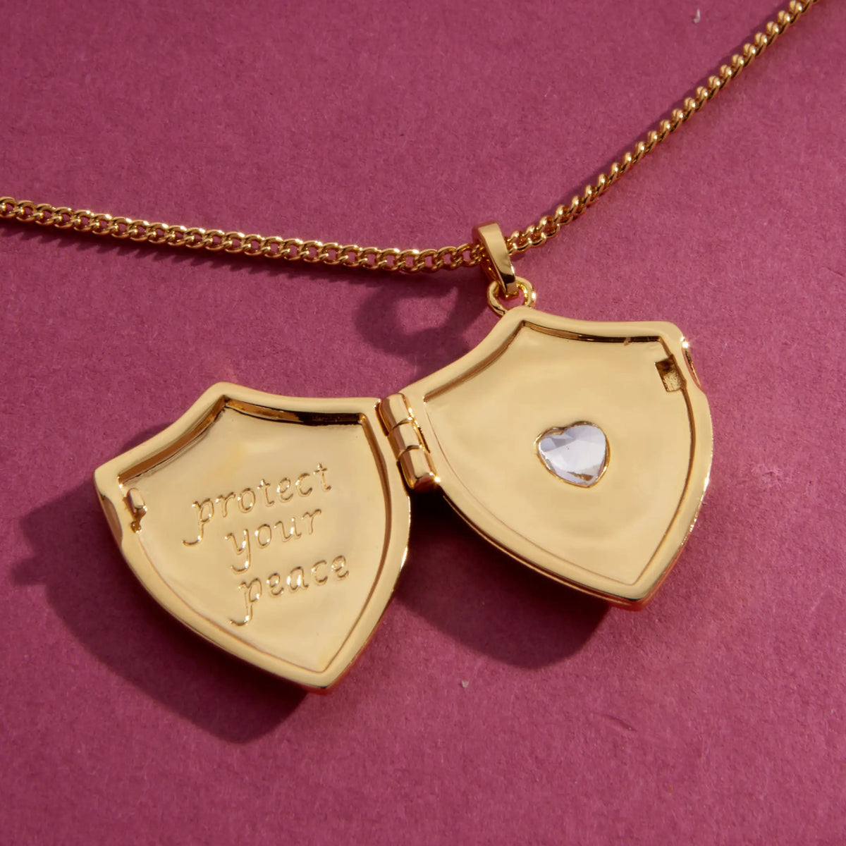 Shield Locket Necklace open, 18k gold-plated shield locket engraved protect your peace with a CZ heart inside, on a gold curb chain, detail on a berry background
