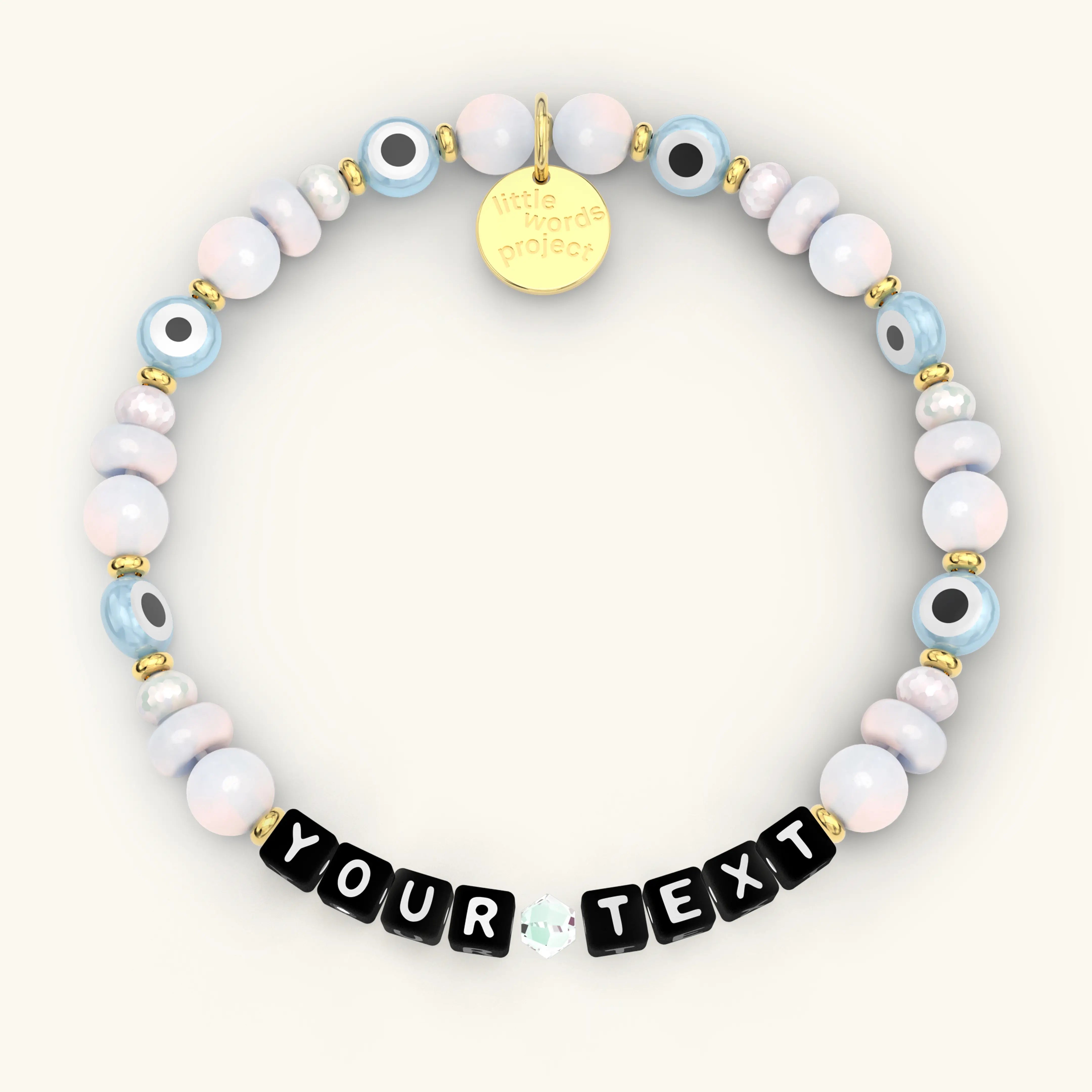 Custom Mystic Blessings beaded bracelet with black letter cubes, pastel pink and blue iridescent beads, blue evil eye beads, and gold hardware, product flat-lay on cream background