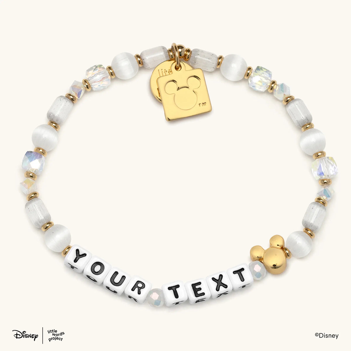 Mickey Custom Magic Maker Disney Mickey & Friends custom bracelet with white letter beads spelling YOUR TEXT, white cat's eye, frosted white, and iridescent clear crystal beads, gold spacers, a gold plated Mickey Mouse bead, and a gold Mickey Mouse logo tag, flat lay