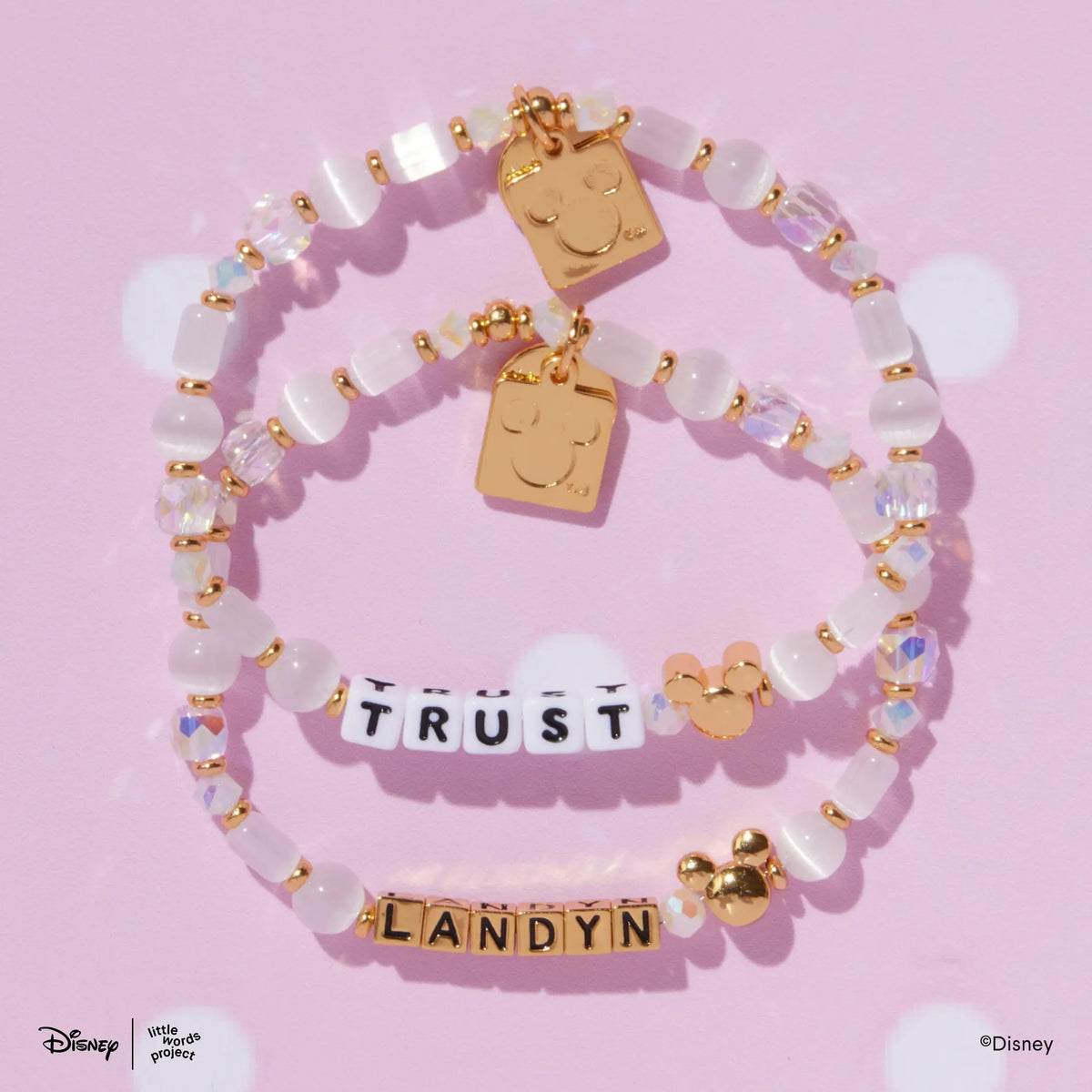 Two Mickey Custom Magic Maker Disney Mickey & Friends custom bracelets with white and gold letter beads, white cat's eye and iridescent crystal beads, flat lay on pink polka dot background