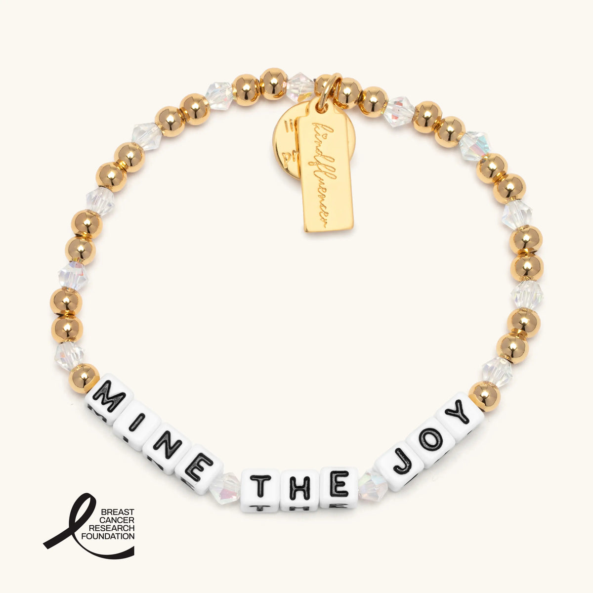 Mine the Joy bracelet in the Golden Hour bead pattern, white letter beads with gold plated beads and iridescent clear crystals, a gold Kindfluencer tag, flat-lay on cream background with the Breast Cancer Research Foundation logo