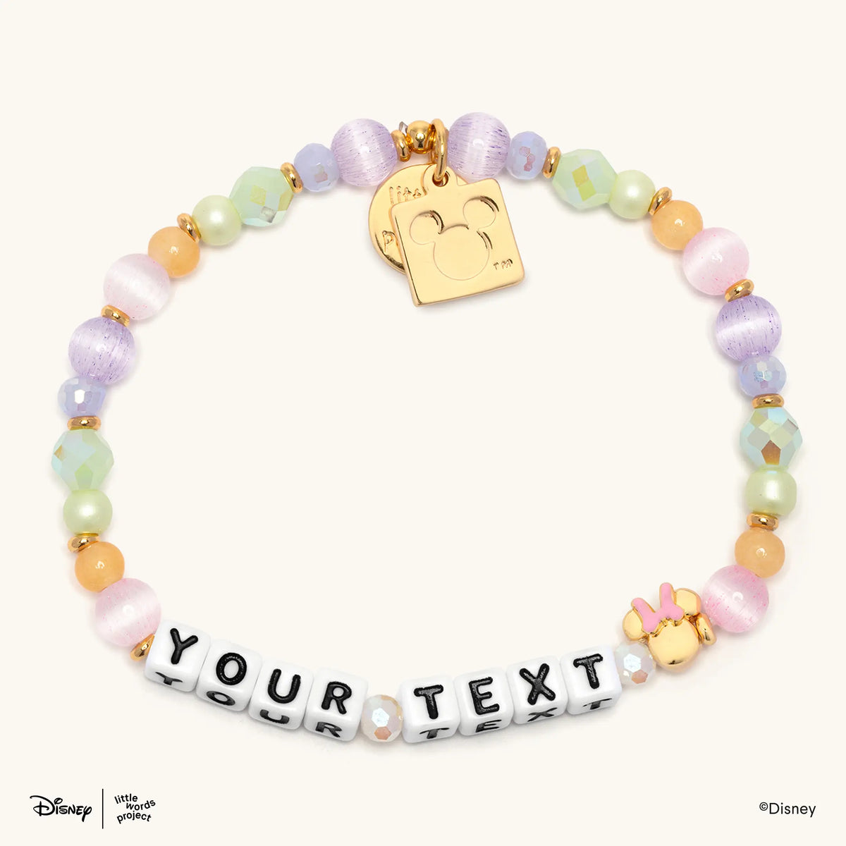 Minnie Custom Dot Darling Disney Mickey & Friends custom bracelet with white letter beads spelling YOUR TEXT, pastel lavender and pink cat's eye beads, mint and lavender iridescent crystals, pale green pearls, peach beads, a gold plated Minnie Mouse bead with a pink bow, and gold Little Words Project and Mickey Mouse logo tags, flat lay