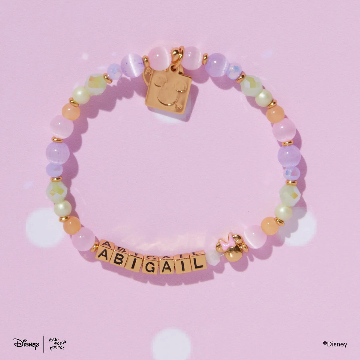 Minnie Custom Dot Darling Disney Mickey & Friends custom bracelet with gold letter beads and a gold plated Minnie Mouse bead with a pink bow, flat lay on pink polka dot background