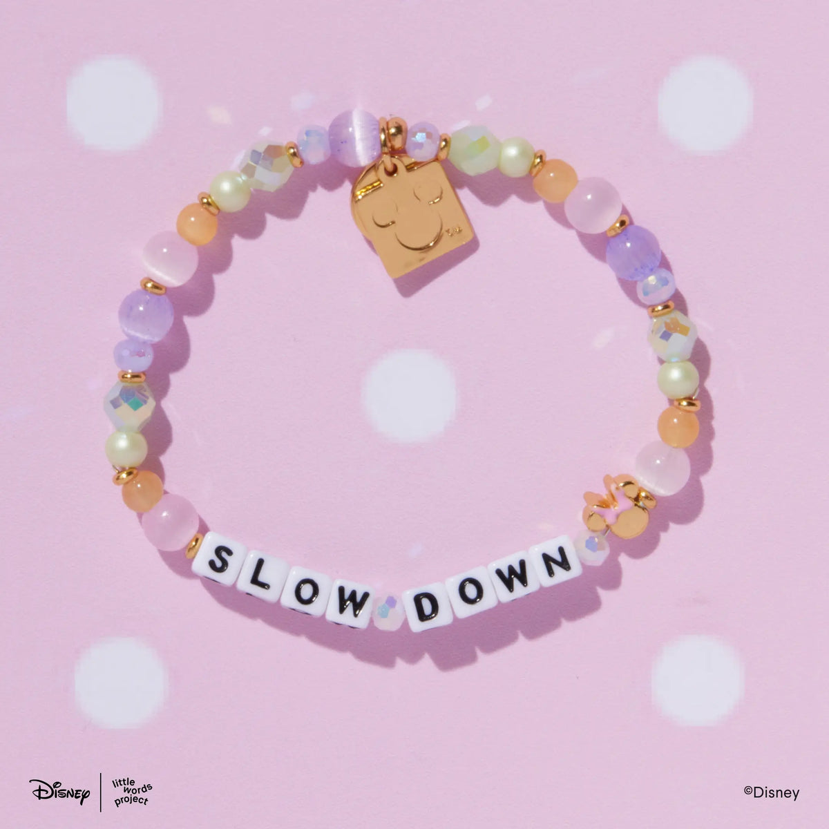 Minnie Custom Dot Darling Disney Mickey & Friends custom bracelet with white letter beads and a gold plated Minnie Mouse bead with a pink bow, flat lay on pink polka dot background