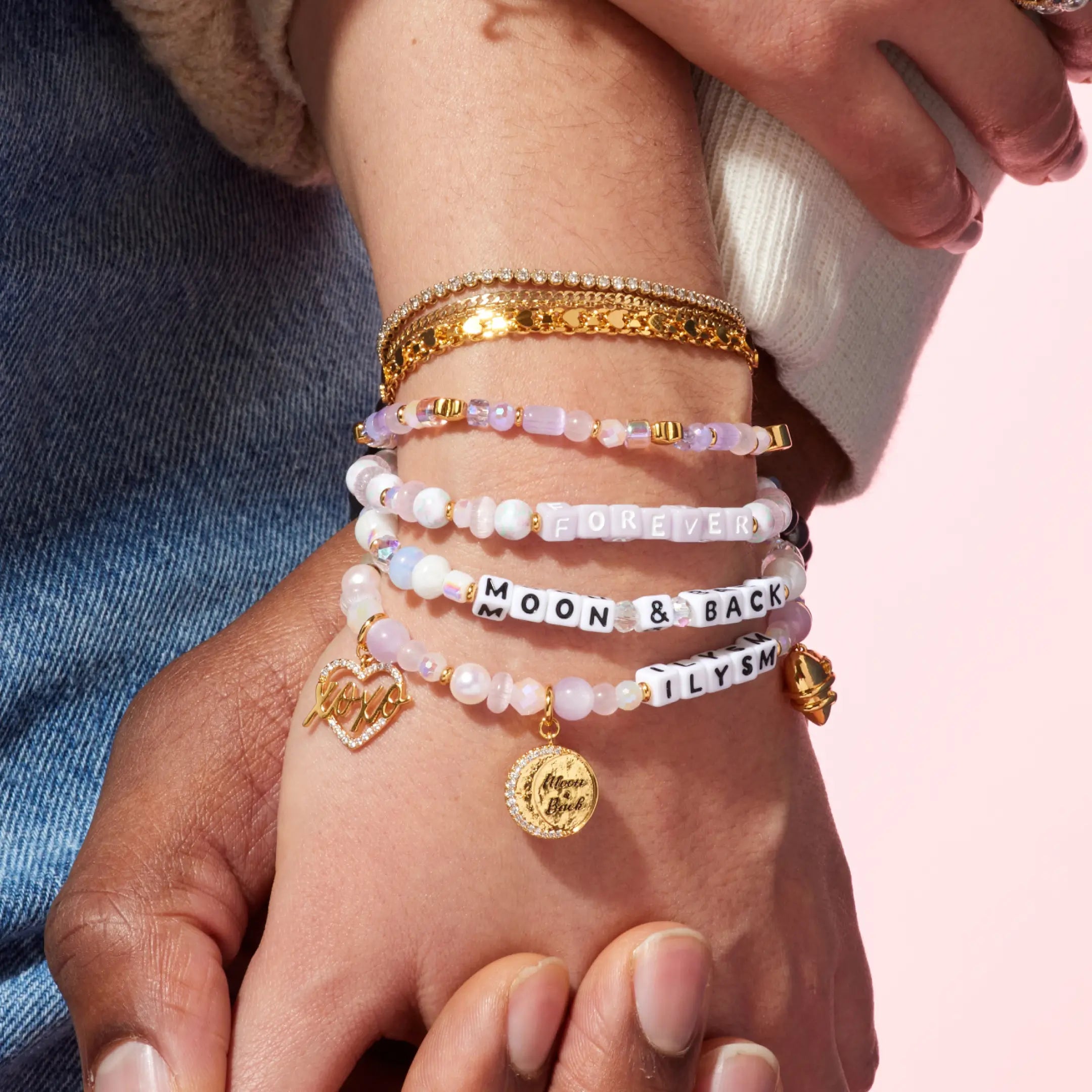 Close-up of a hand wearing multiple bracelets with various charms and text on a pink background
