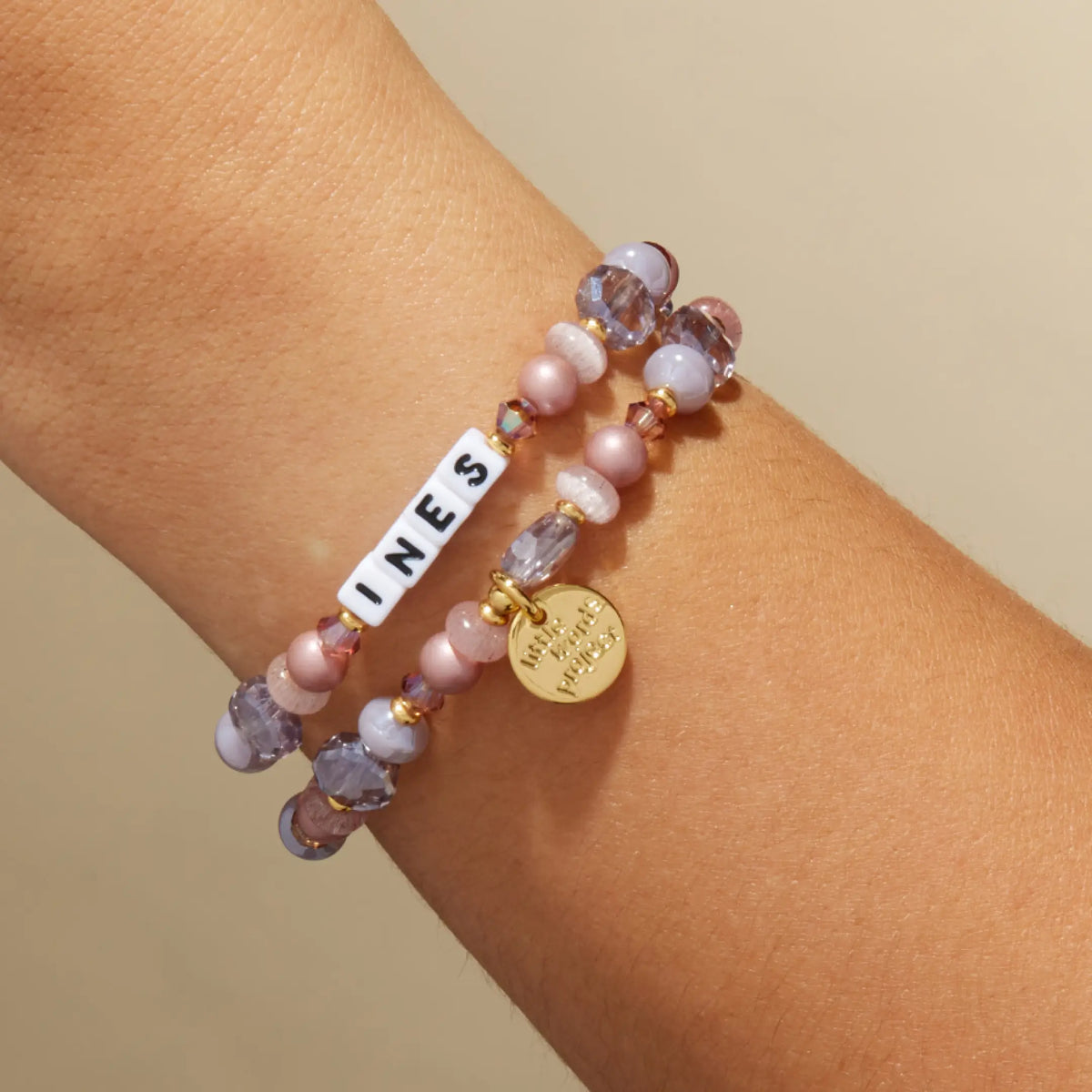 Custom Nice Spice bracelet with white letter beads reading Ines, mauve pearl and lavender crystal beads with a gold LWP charm, on-model on wrist against a tan background