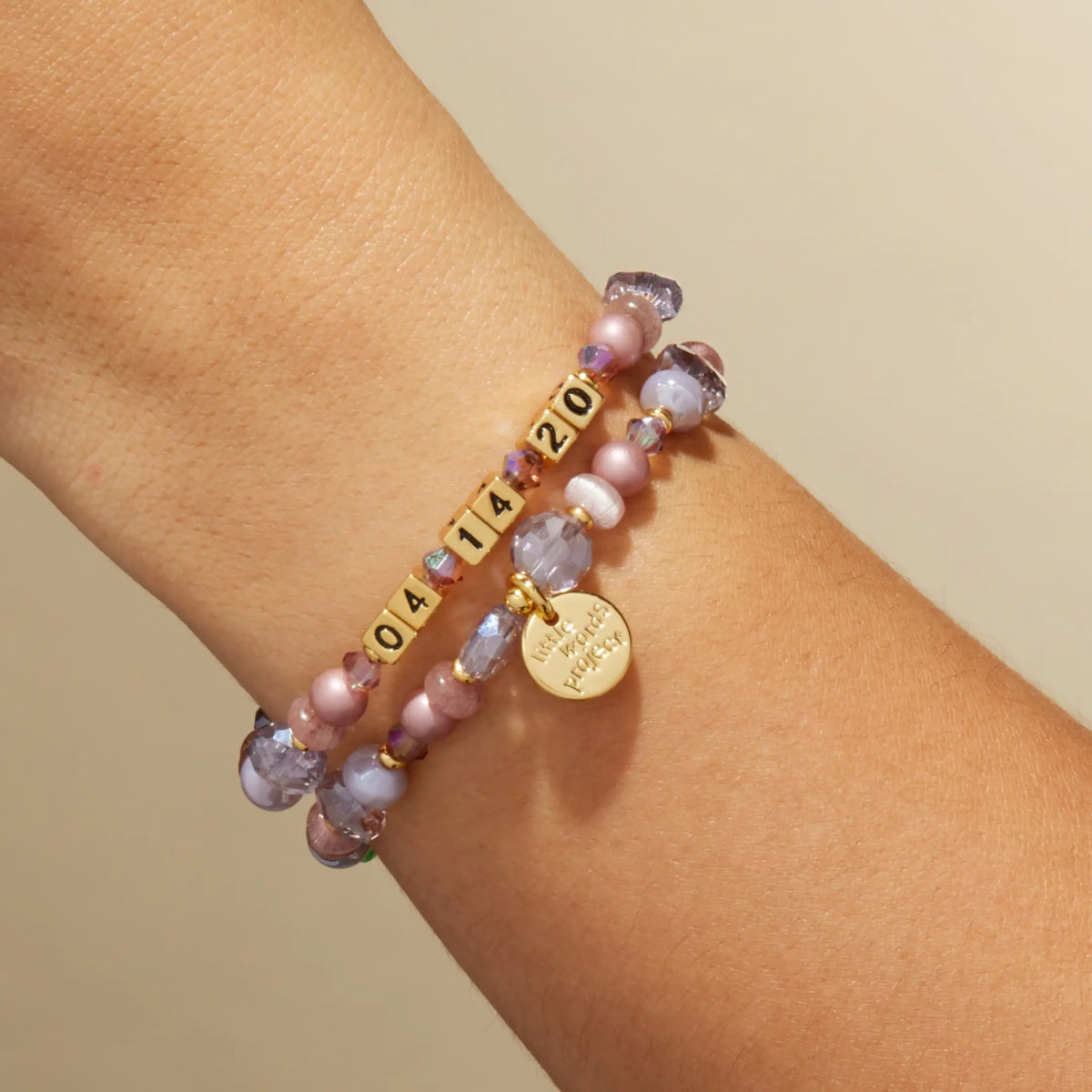Custom Nice Spice bracelet with gold letter beads reading 04 14 20, mauve pearl and lavender crystal beads with a gold LWP charm, on-model on wrist against a tan background